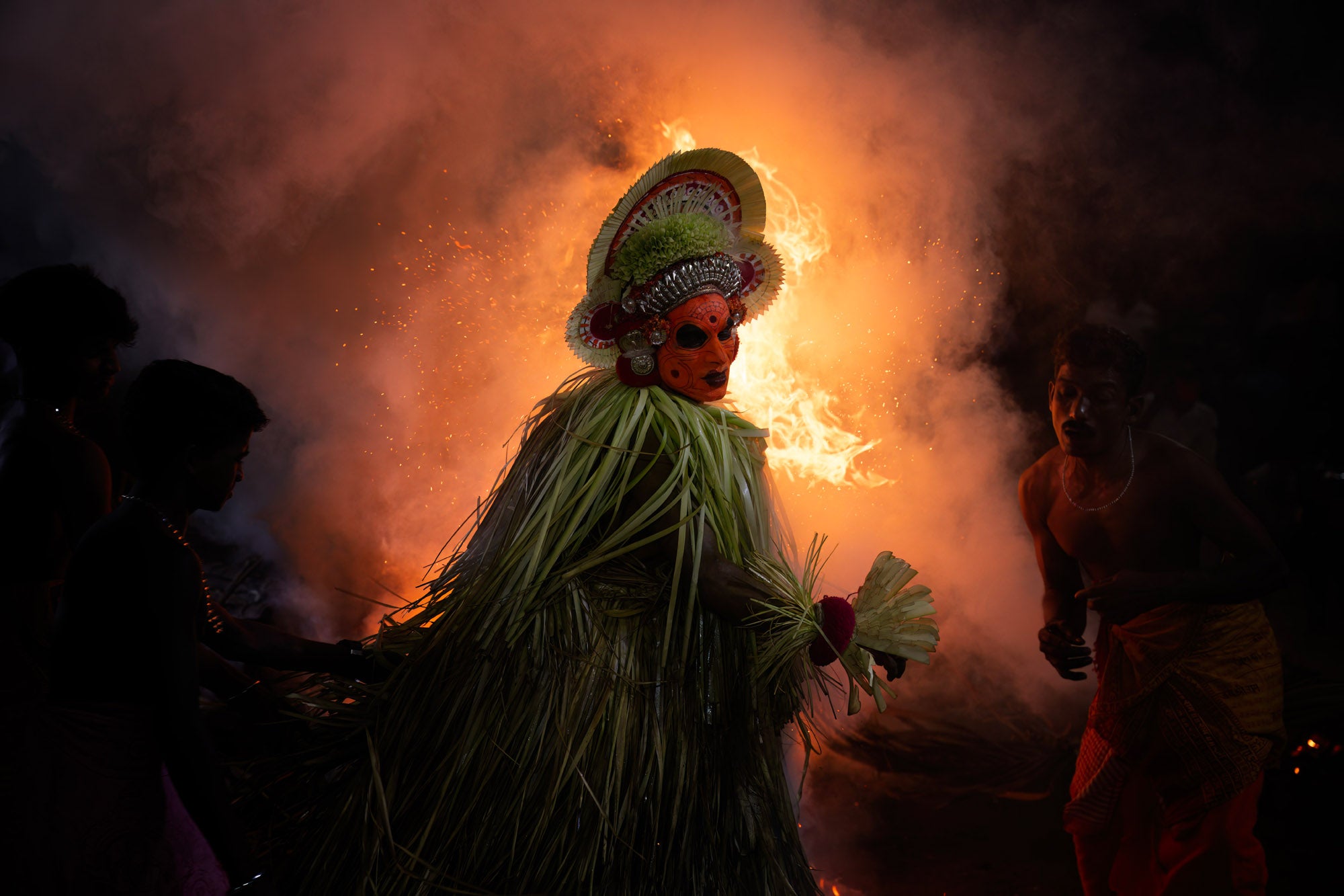 A person dressed with a headpiece and painted face moves in front of a fire. 