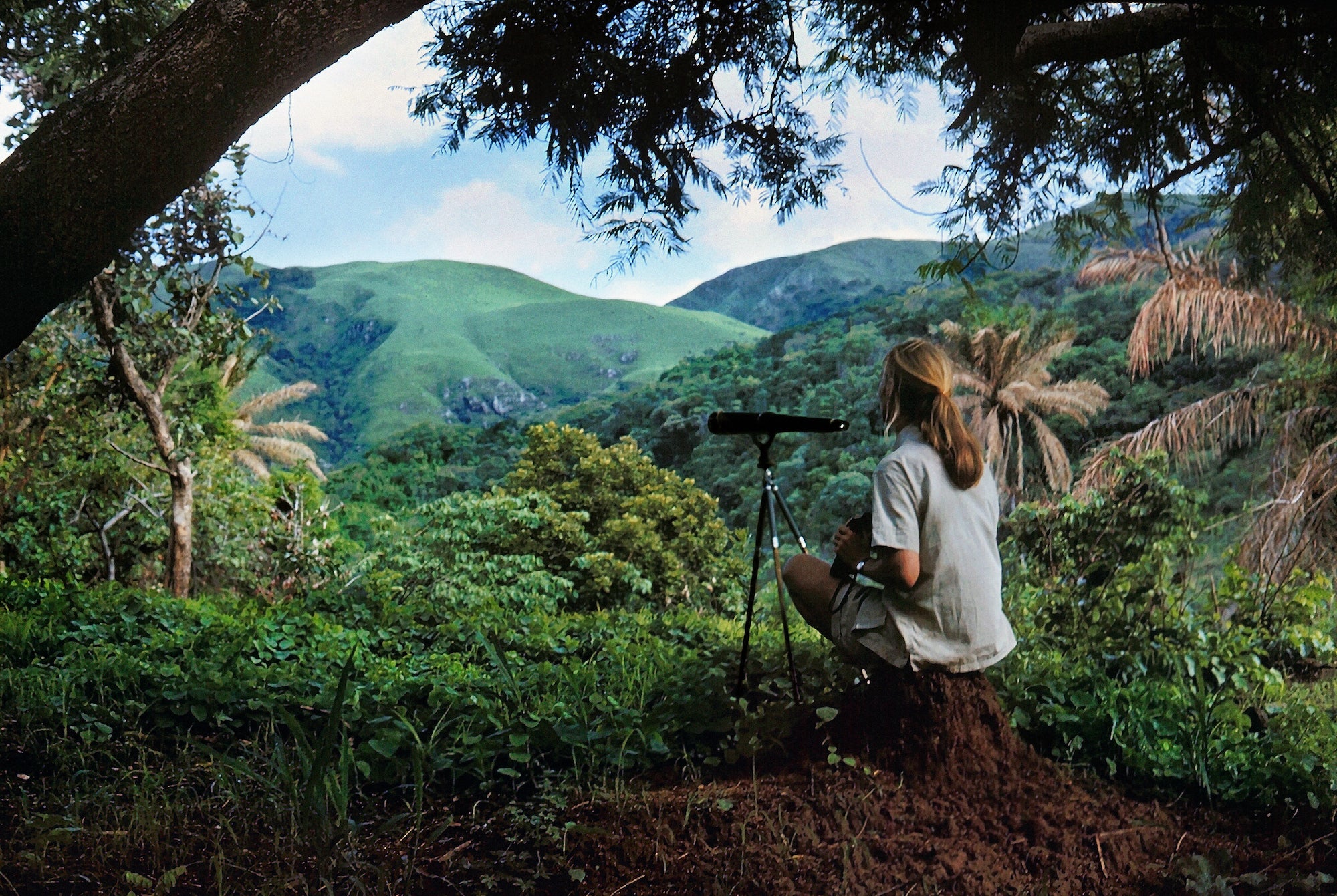 a woman sits on a hill top with binoculars and a telescope