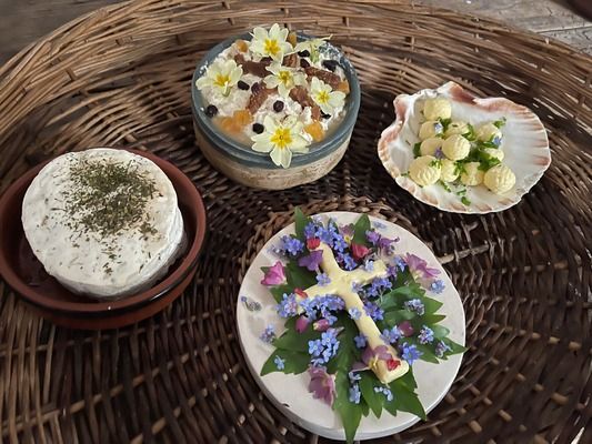 various easter cheese and butters are on a table. a cross made from butter is laid on a bed of greens with purple flowers. a round cheese is sprinkled with greens. a dish with wet cheese is decorated with yellow and white flowres. a scallop bowl of butter balls is decorated with greens.