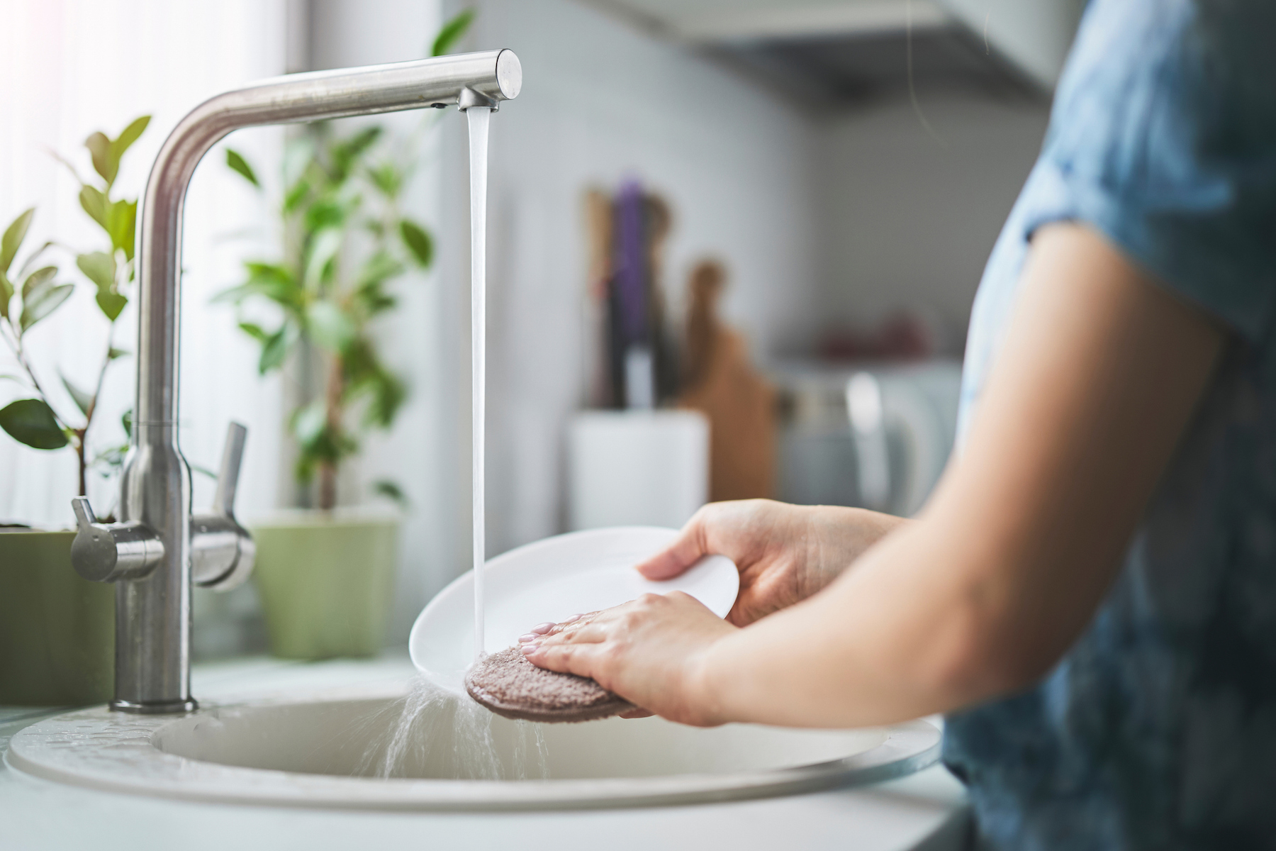 A person cleaning dishes under running water in a sunny kitchen, emphasizing cleanliness and eco-friendly habits. Potted plants complement the refreshing and organized setting