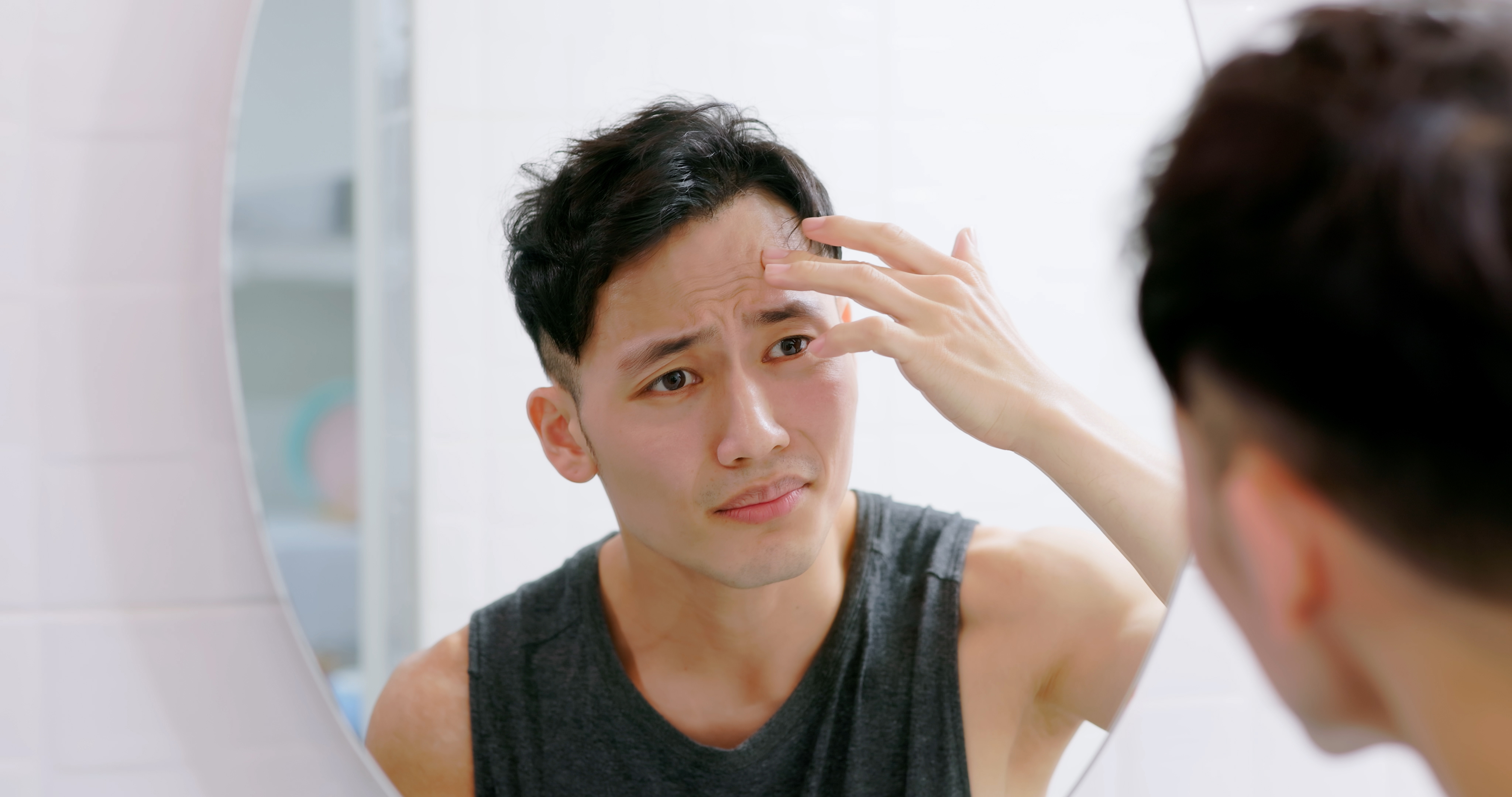 A young man with short dark hair examines his forehead closely in the bathroom mirror, lightly touching his skin and frowning at visible lines.