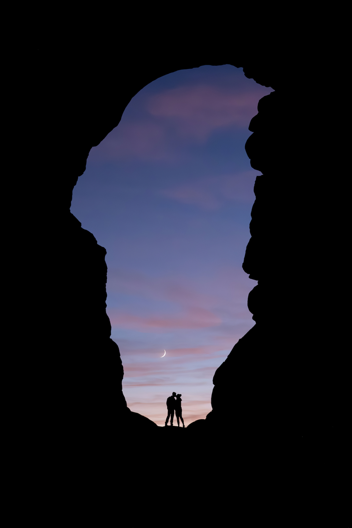 a couple kisses in the hole formed by rock formations. a crescent moon in the background