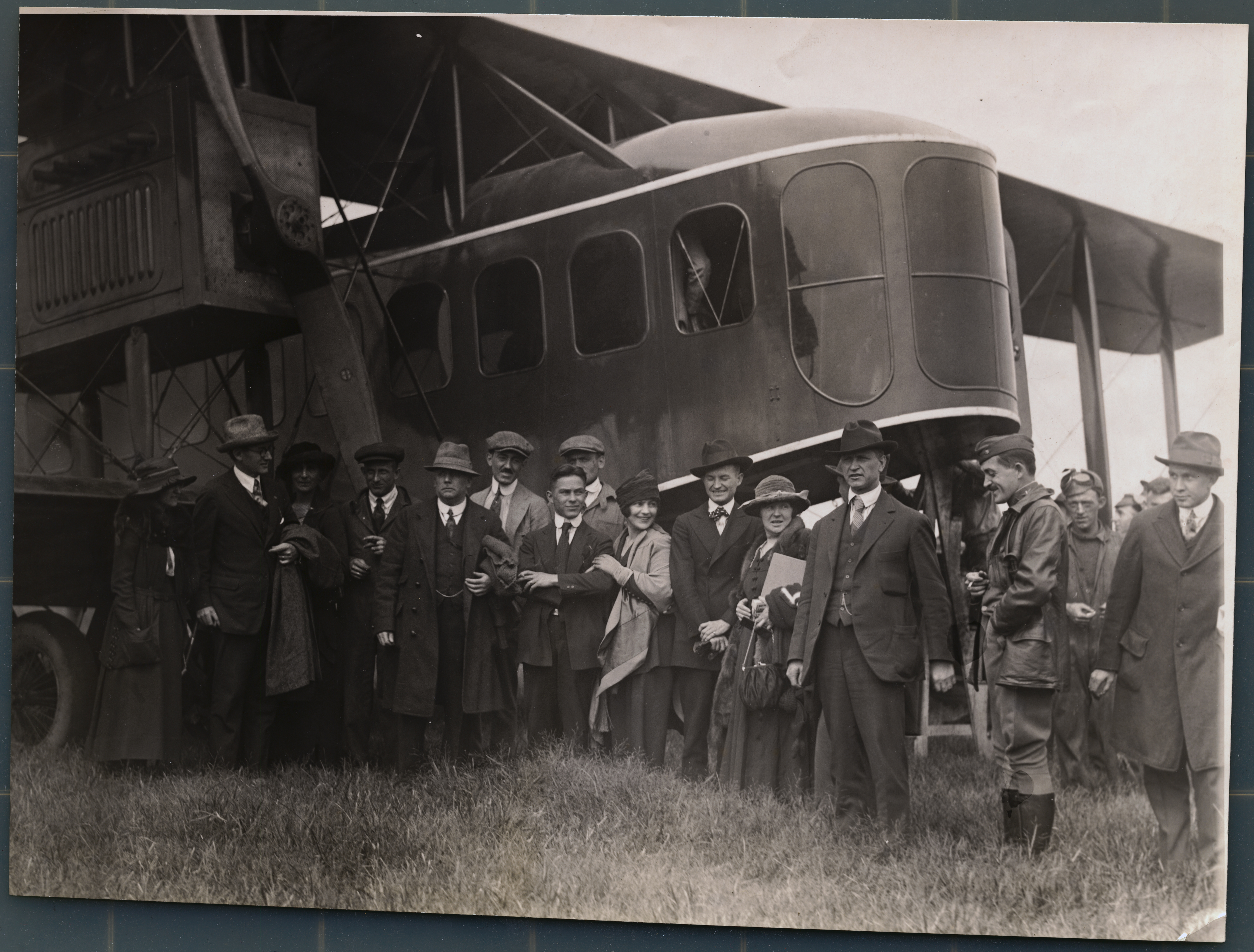 Black‑and‑white photograph of a group of early 20th‑century air passengers gathered on an airfield, standing in front of a large biplane. The men and women—dressed in period-appropriate coats, hats, and scarves—pose or move casually near the aircraft’s fuselage and engine. 