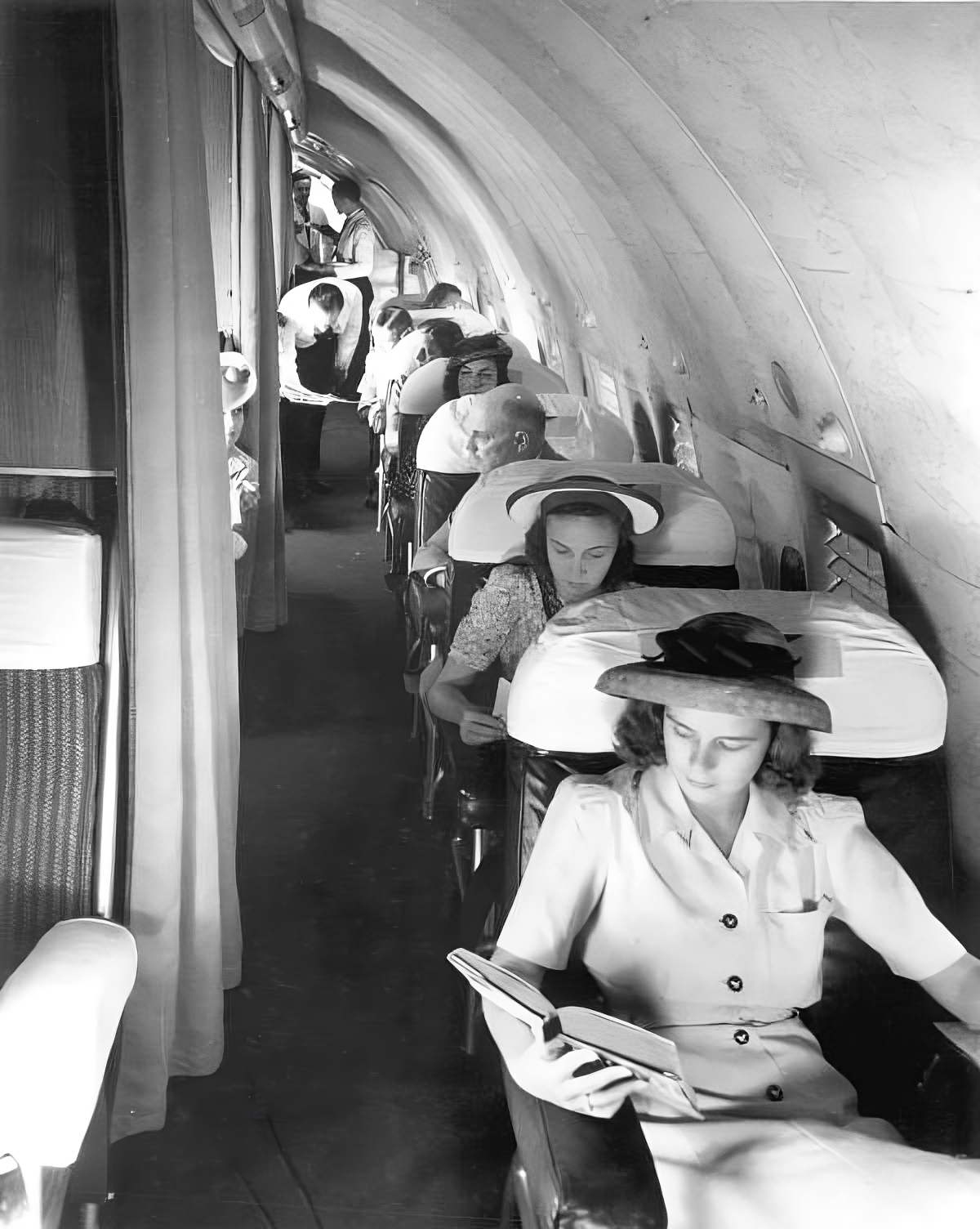 A black and white image shows the interior of a vintage airplane. Seated closest to the camera, a young female passenger in a hat reads a book. Behind her another woman in a hat reads.