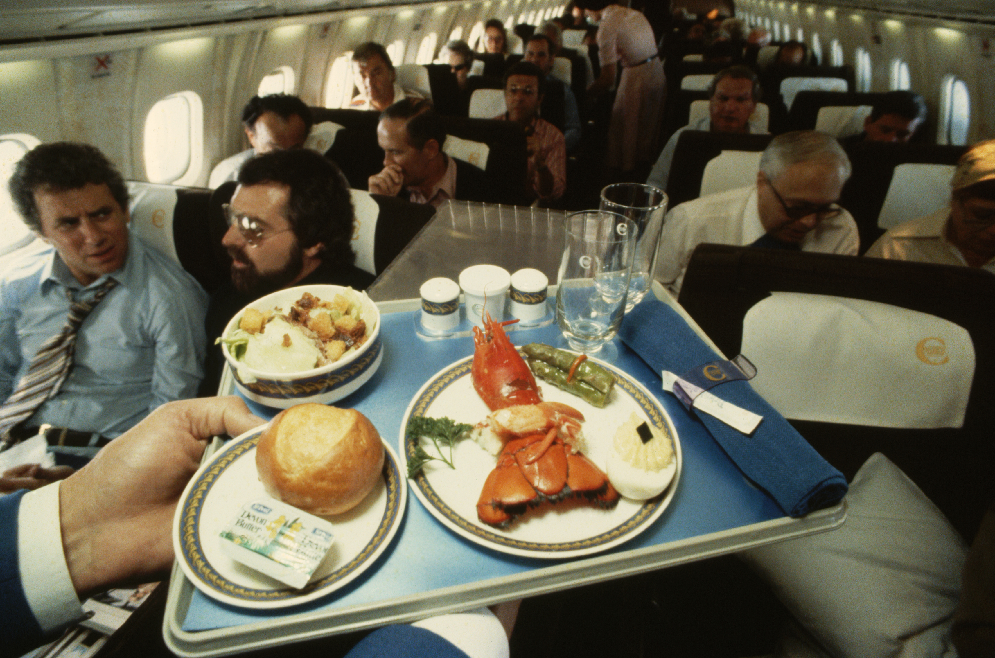 A tray of luxurious in-flight food aboard the Concorde. The meal includes lobster, asparagus, a hard-boiled egg, a dinner roll, a small Caesar salad, salt and pepper shakers, two empty cups, and a rolled napkin. Behind the tray, the narrow cabin of the Concorde is visible, with passengers seated two-by-two on either side of the aisle. All the passengers appear to be men, dressed in business attire. Closest to the tray is a man in a blue shirt and tie, seated and looking forward. The image evokes the exclusivity and refinement of supersonic air travel.