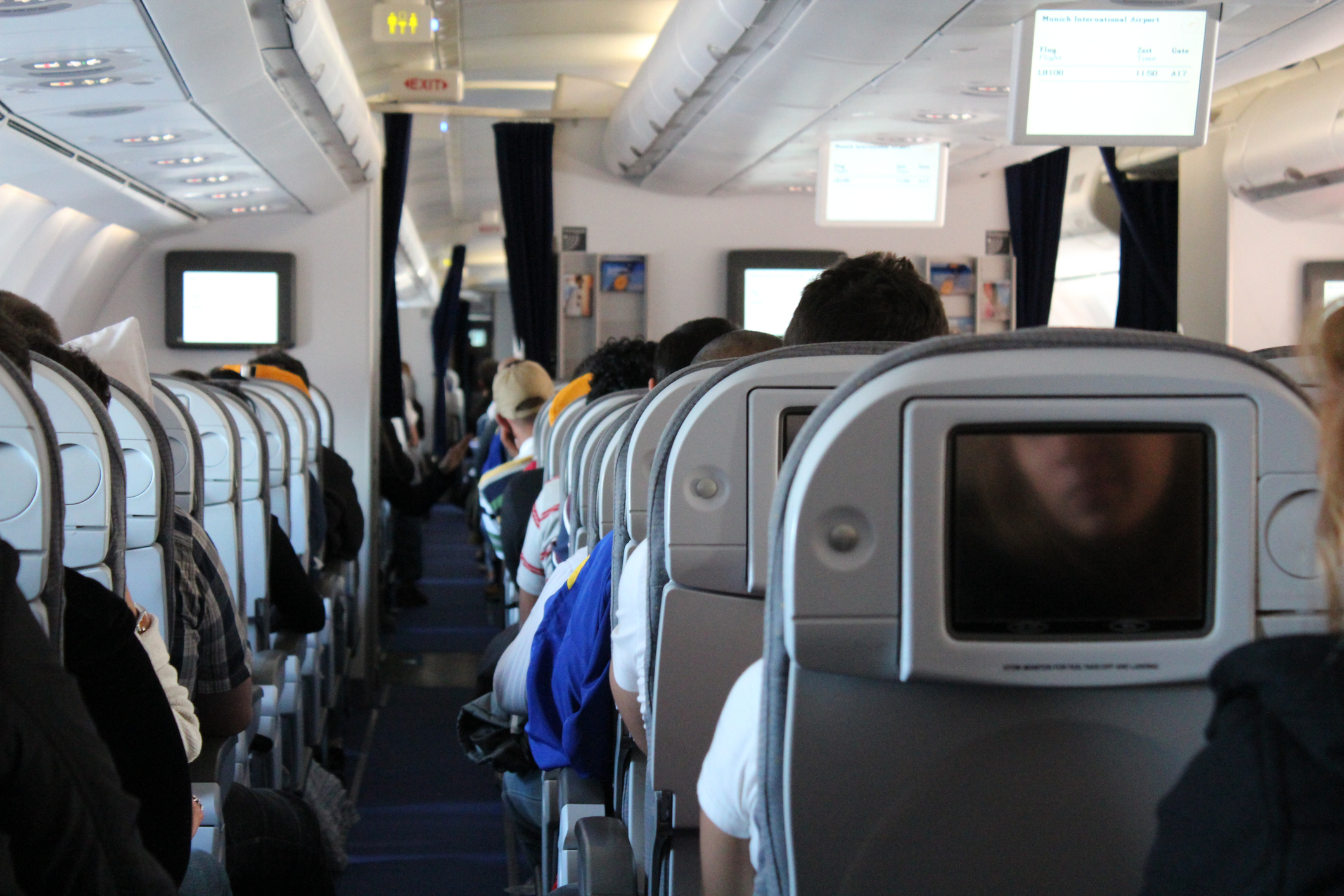 View from the back of an airplane cabin showing the backs of passengers’ heads as they sit quietly in their seats, facing forward and waiting for takeoff. The aisle runs down the center-left of the image, flanked by overhead luggage bins. Seatback monitors are visible. At the front of the cabin, an illuminated exit sign and bathroom sign can be seen. The scene captures a typical pre-flight moment inside a commercial aircraft.