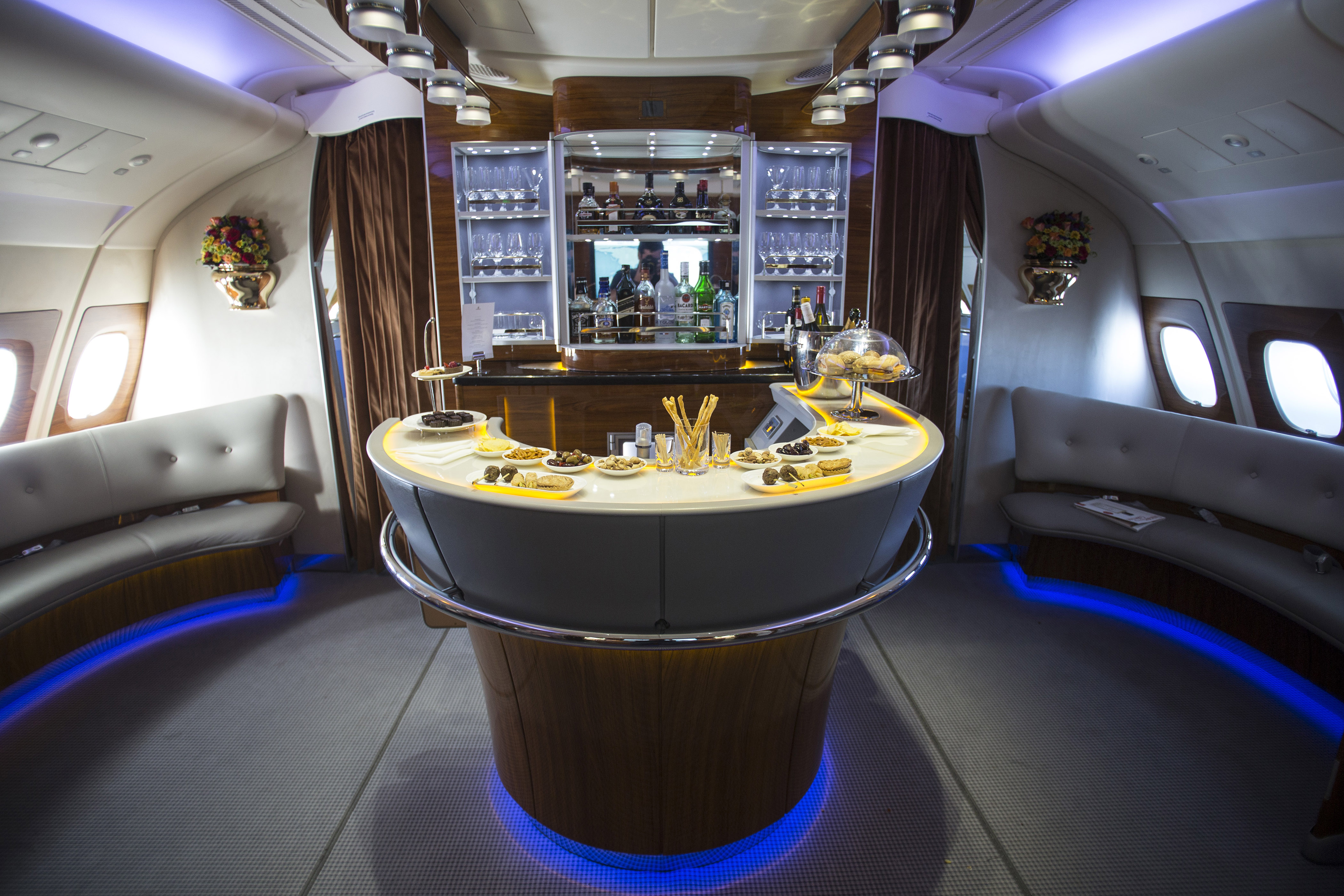 A semicircular bar area inside an Emirates airplane cabin, featuring a variety of snacks such as breadsticks, sandwiches, and other bite-sized foods arranged on the counter. Surrounding the bar is curved seating. Behind the bar, shelves hold glasses and bottles of alcohol, creating a sleek and inviting onboard lounge space.