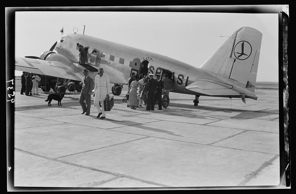 A black-and-white photograph showing a group of airline passengers beside the open door of a LOT Polish Airlines aircraft on the tarmac at Lydda (now Lod) Airport in Israel. A mix of men and women, dressed in period-appropriate attire—coats, hats, and long dresses—are stepping down the aircraft’s boarding stairs onto the airfield.