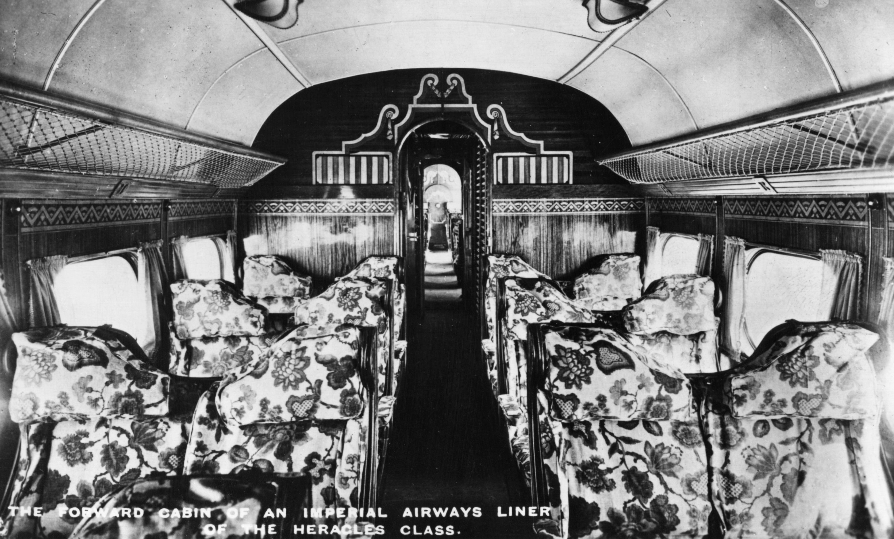 Black-and-white photograph of the passenger cabin aboard an Imperial Airways Handley Page HP42E 'Heracles' airliner, circa 1935. The cabin exudes a Pullman-style elegance, featuring plush upholstered seats with floral fabric arranged in rows along a central aisle. Inlaid wood decorates the walls. Wide windows line the walls, allowing light to softly illuminate the interior, giving a sense of early luxury in air travel. 