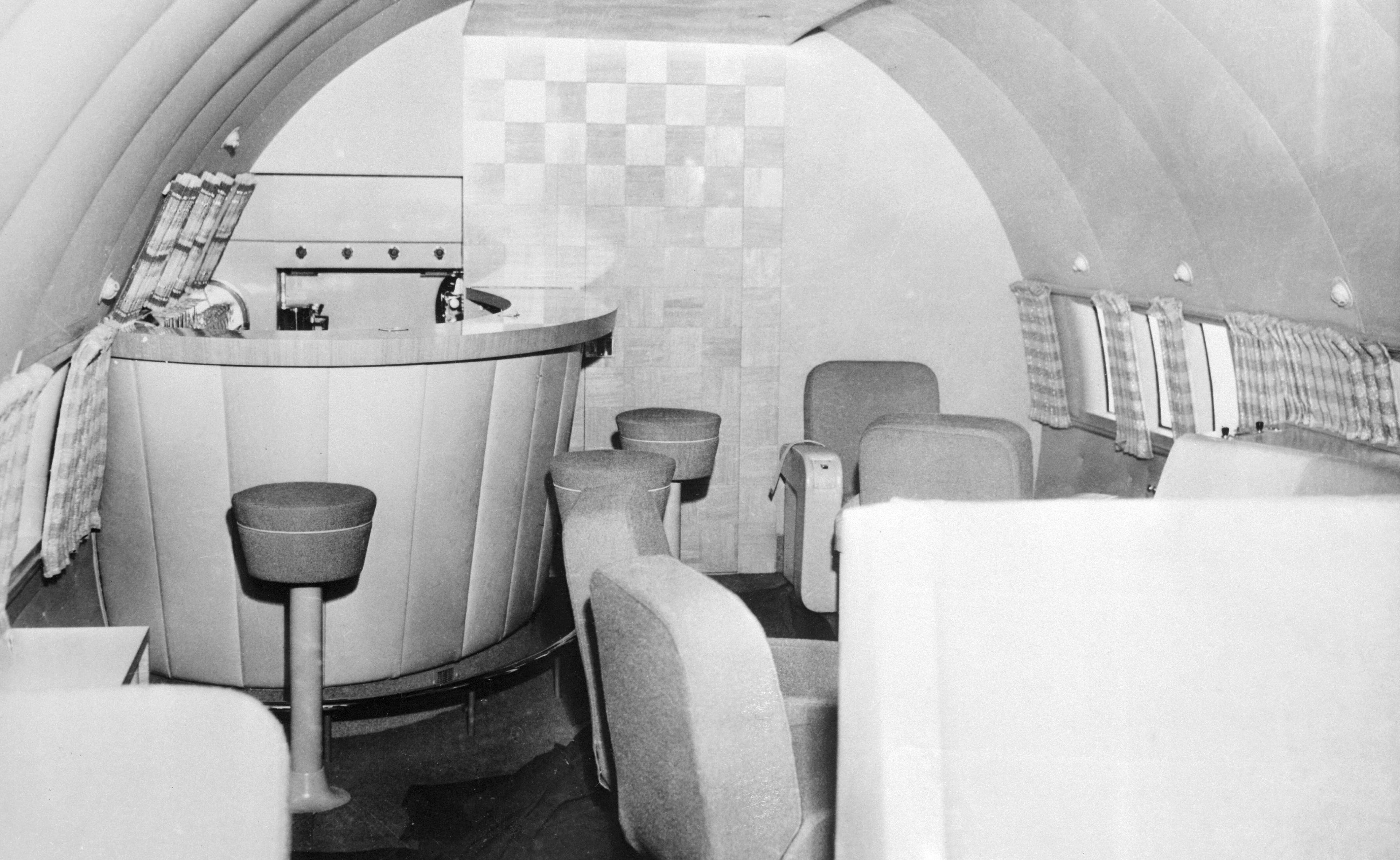 A black-and-white interior photograph of an onboard plane bar fitted with a curved service counter surrounded by three stools. Plush chairs are arranged nearby, although largely out of frame.
