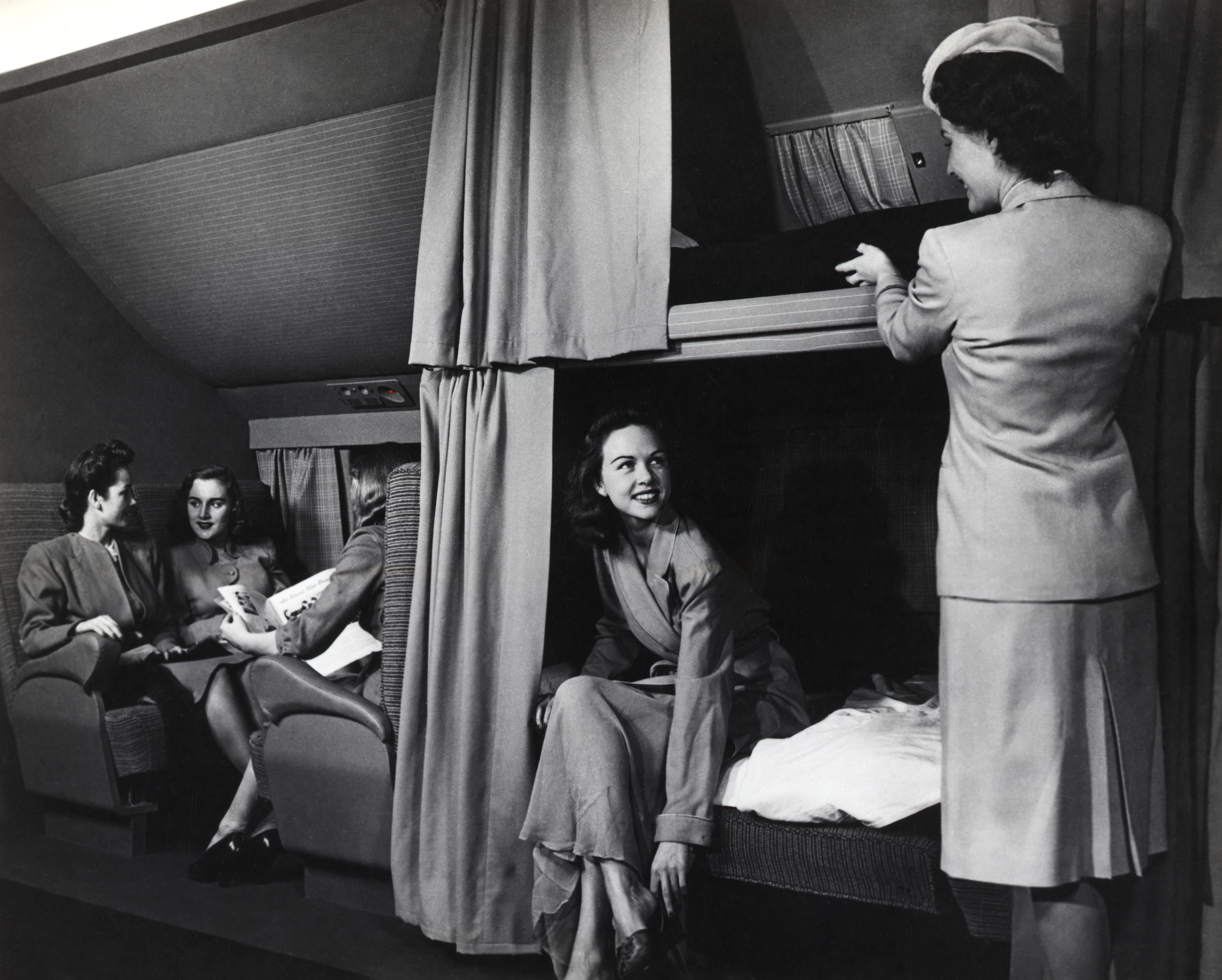Black-and-white photo of a mid-20th-century commercial airplane cabin, likely a Douglas DC-6, featuring a sleeping compartment. On the left, three women dressed in tailored suits sit and chat in upholstered seats; one holds a magazine. In the center, another woman smiles while seated on the edge of a lower bunk bed, adjusting her shoe. On the right, a flight attendant in a uniform and cap demonstrates the upper berth, lifting the bedding. Curtains frame the bunks, and the space exudes the elegance and comfort of postwar luxury air travel in the 1940s or 1950s.