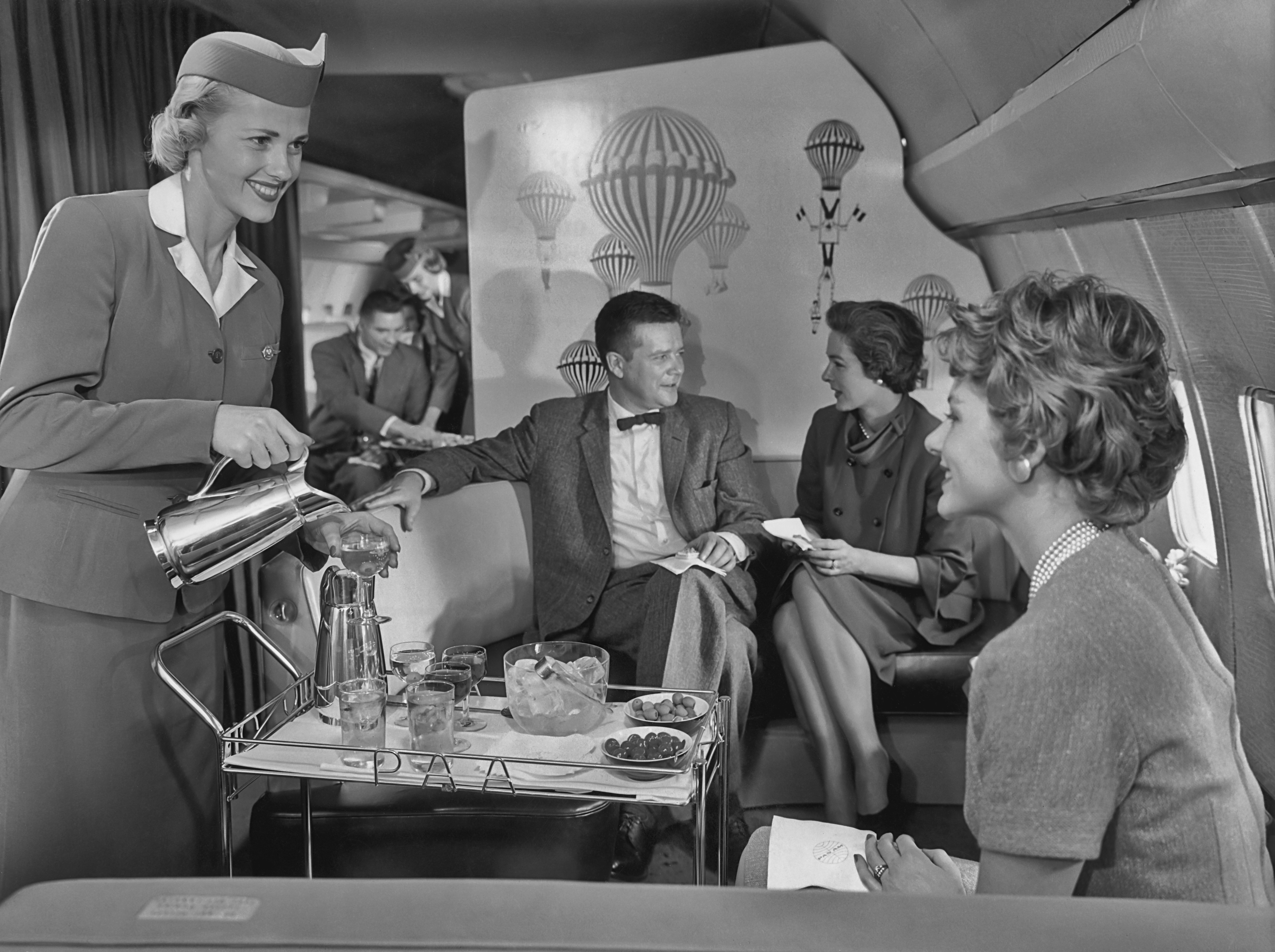 Black-and-white photo of a stylish mid-century airplane lounge aboard a Pan American World Airways (PAA) flight, circa 1958. A smiling flight attendant in a tailored uniform pours drinks from a metal pitcher onto a tray table stocked with ice, glassware, and snacks. In the background, well-dressed passengers—two women and a man in a suit and bow tie—chat on a curved couch in front of a decorative panel featuring whimsical hot air balloons. Another attendant serves a tray to a man in the rear. The setting exudes the glamour and social atmosphere of luxury air travel during the golden age of commercial aviation.