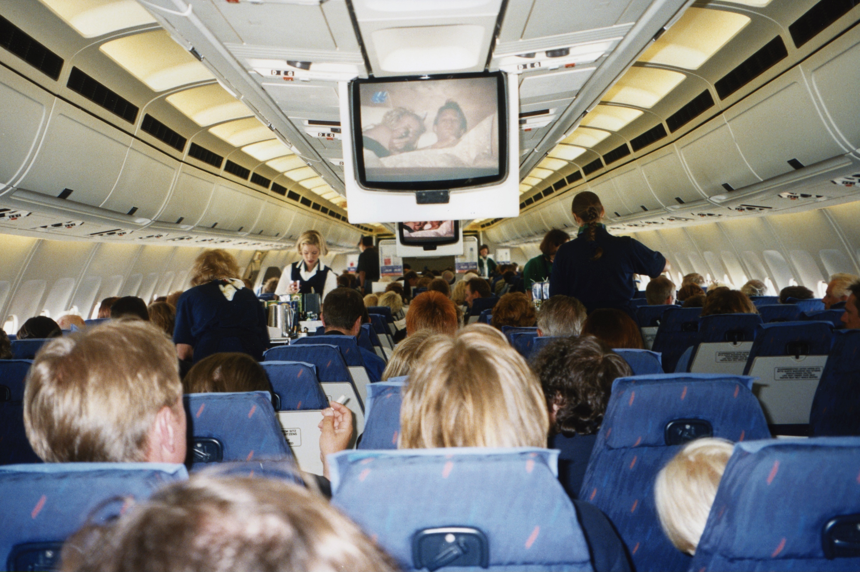 The interior of a crowded airplane with passengers seated closely together. On both sides, flight attendants are pouring drinks for the passengers. Some passengers are smoking, and a small TV monitor has descended from the ceiling, showing a movie that the passengers are watching. The overall atmosphere is lively and nostalgic, reflecting a bygone era of air travel.