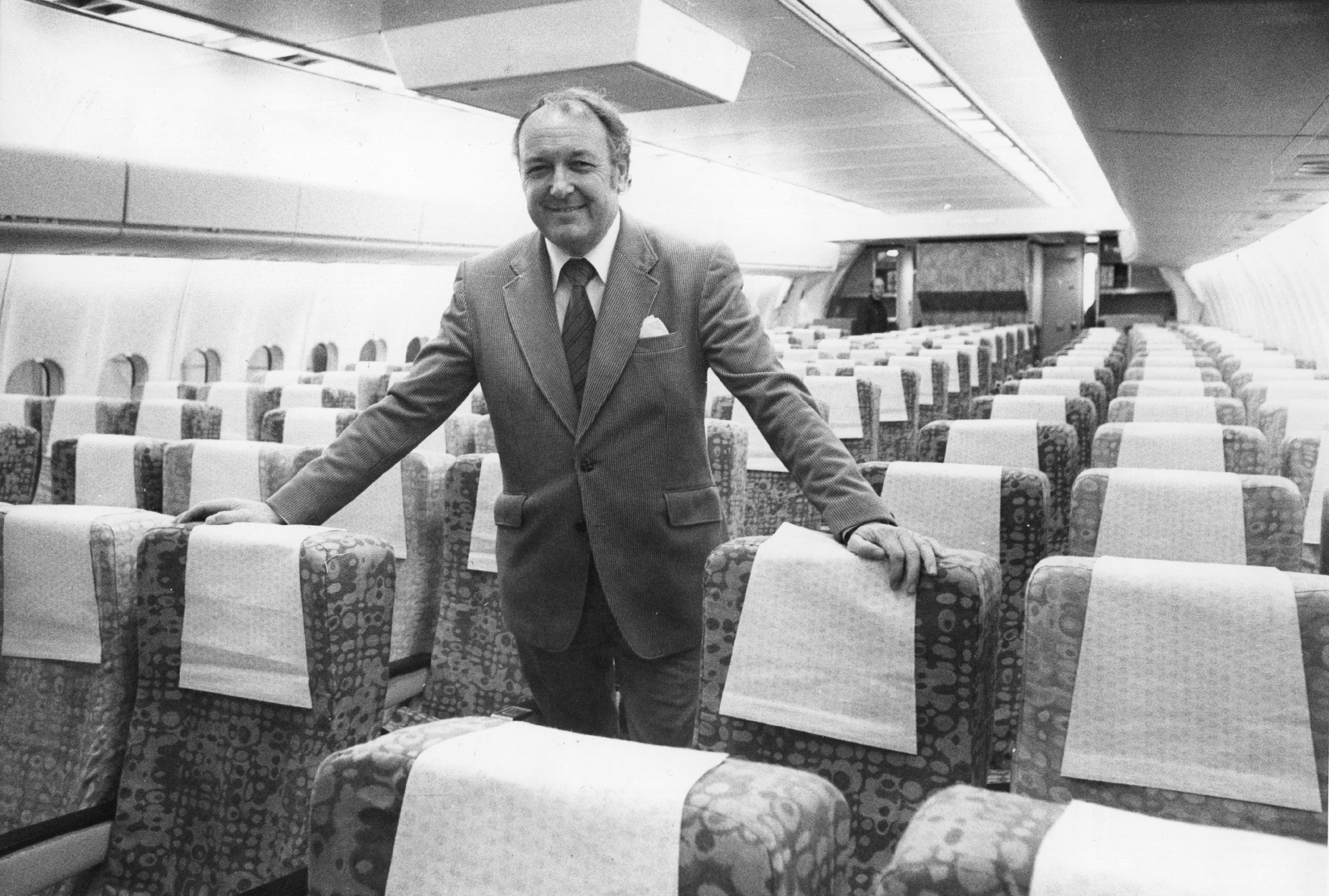 A smiling middle-aged man, identified as airline pioneer Freddy Laker, stands in an empty airplane cabin with closely spaced seats. Slightly balding, he gazes toward the camera, embodying the no-frills, accessible air travel he helped popularize. The sparse cabin layout highlights the utilitarian design of early budget airlines. The airplane seats have a splotchy pattern.