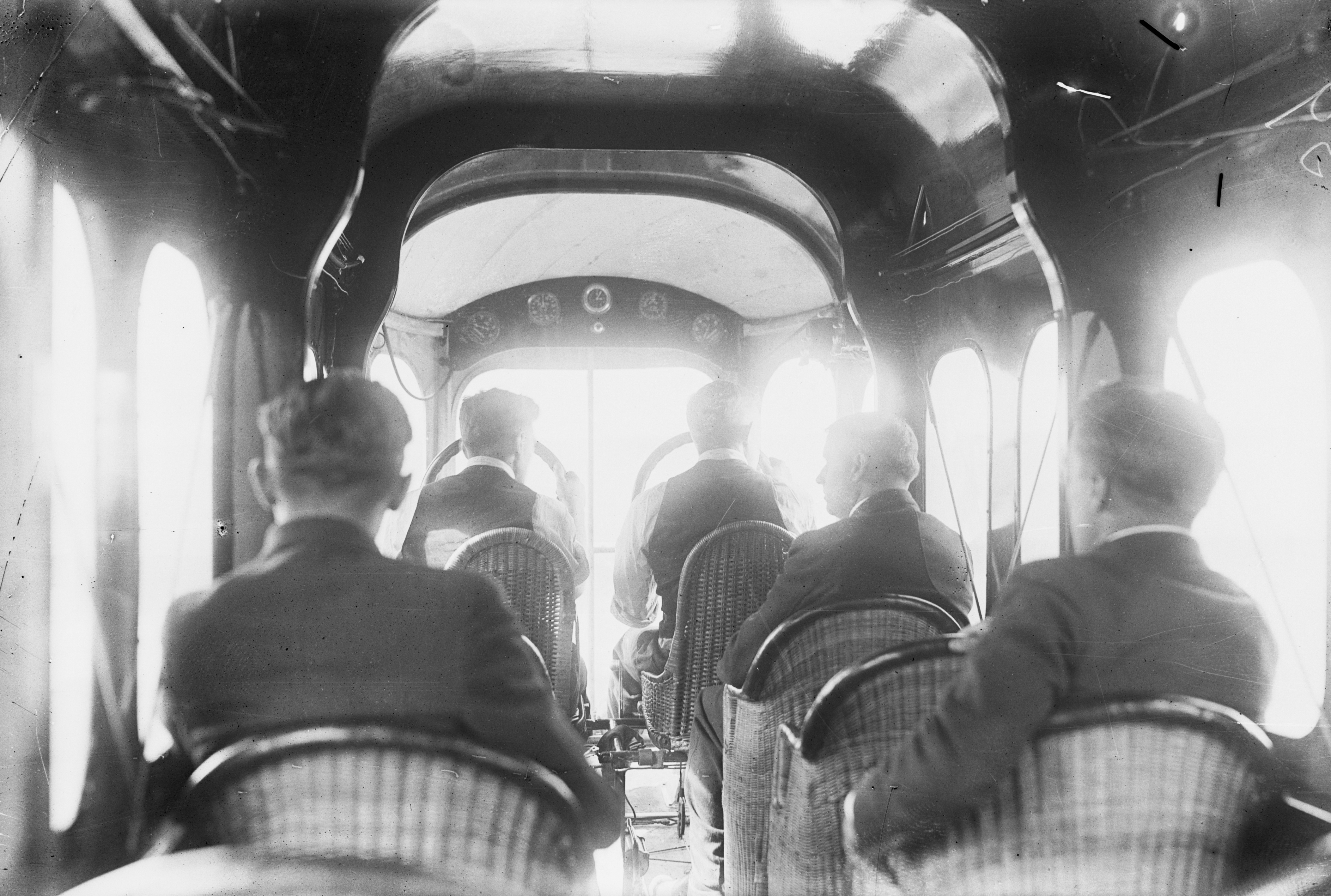 Black-and-white interior view of an early passenger airplane cabin. The cabin appears boxy with bare walls. A few passengers are seated on freestanding cane‑backed chairs arranged along the cabin, facing forward. Light filters in through small windows along the fuselage. The airplane’s structural elements—such as ribs and spars—are subtly visible behind the walls, giving a utilitarian feel. Overall, the scene evokes the early days of commercial flight.