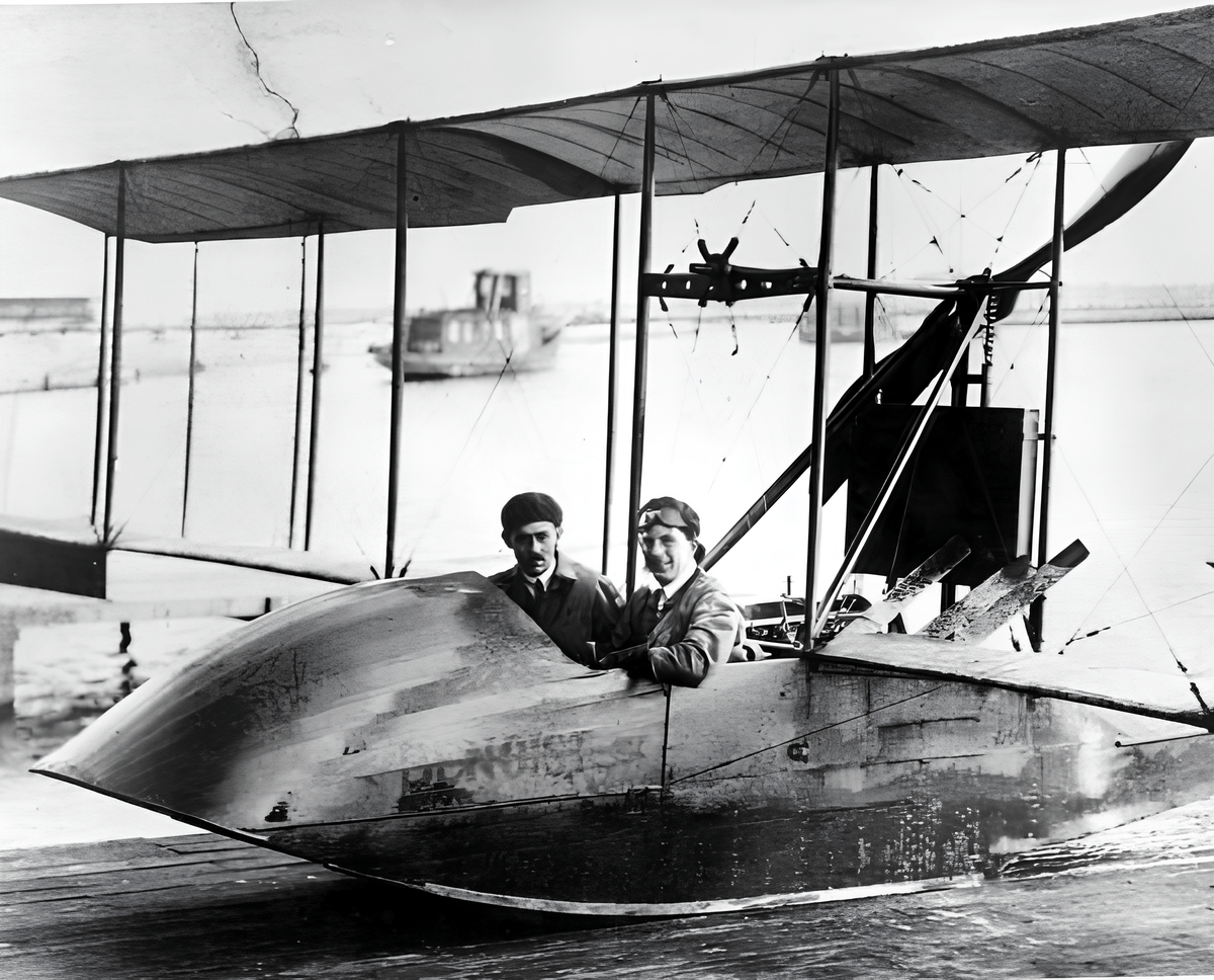 Black-and-white historical photograph of two men seated in the open cockpit of a biplane seaplane docked at the water’s edge. The man on the left wears a dark cap and a suit with a tie, while the man on the right wears a lighter cap and jacket. Behind them, the aircraft’s large fabric-covered wings are supported by struts and wires. In the background, calm water extends to a distant breakwater and a tugboat. 