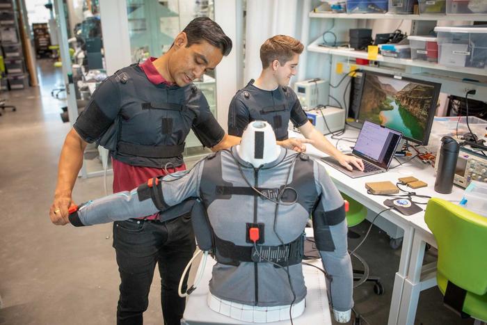 two students working in a lab with a gray vest. the vest has wires and sensors on it that can track movements