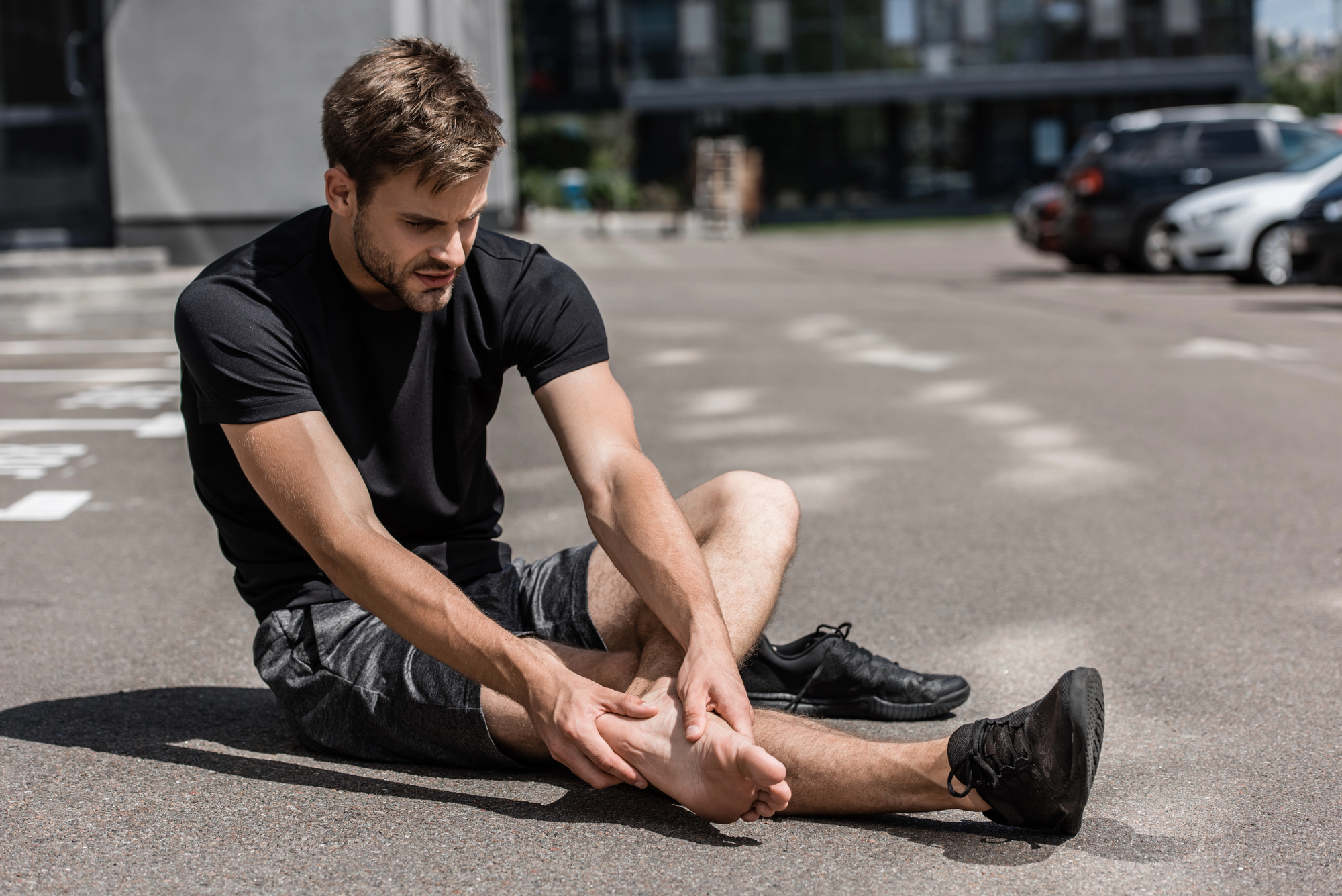 An athletic man wearing a black T-shirt and grey shorts holds his bare left foot in pain. He sits on the pavement in parking lot. His left black running shoe is on the ground next to him. His right foot still has a black shoe on. 