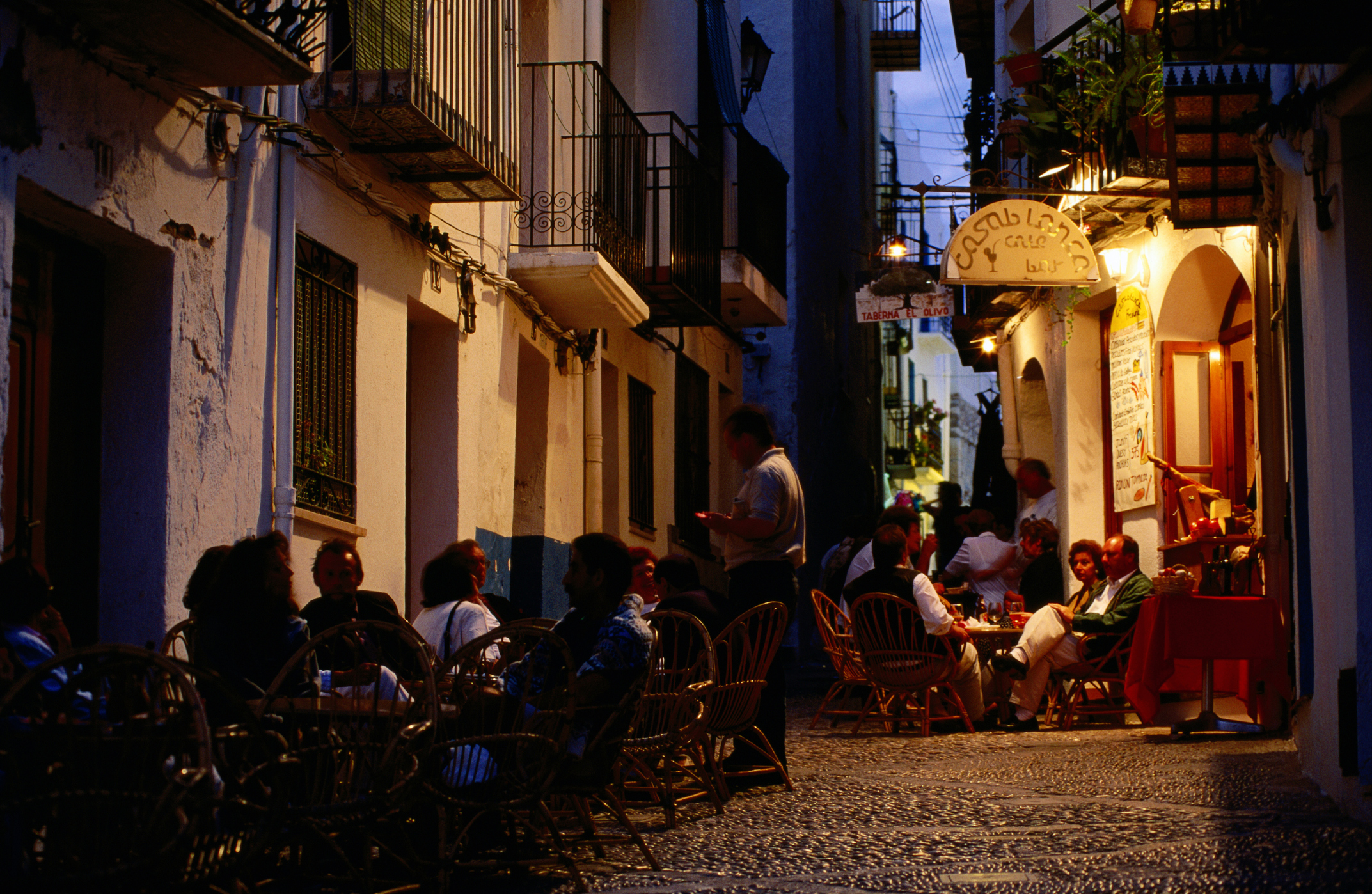 A narrow, cobbled European street at dusk or night, lined with buildings and bustling with people seated at outdoor tables. Warm light spills from open doorways and windows of cafes and restaurants, where groups of people are enjoying meals and conversation. Balconies with railings and potted plants are visible on the upper floors of the buildings.