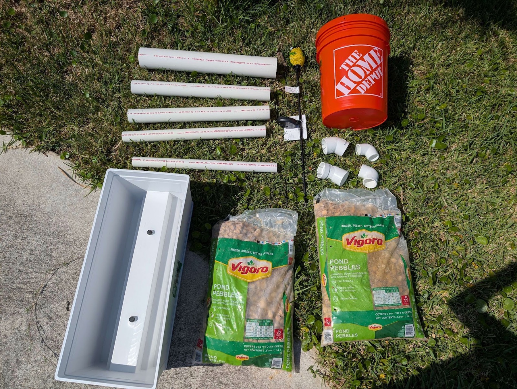 materials laying on the grass: a bucket, pipes, pebbles, a planter, a frog light