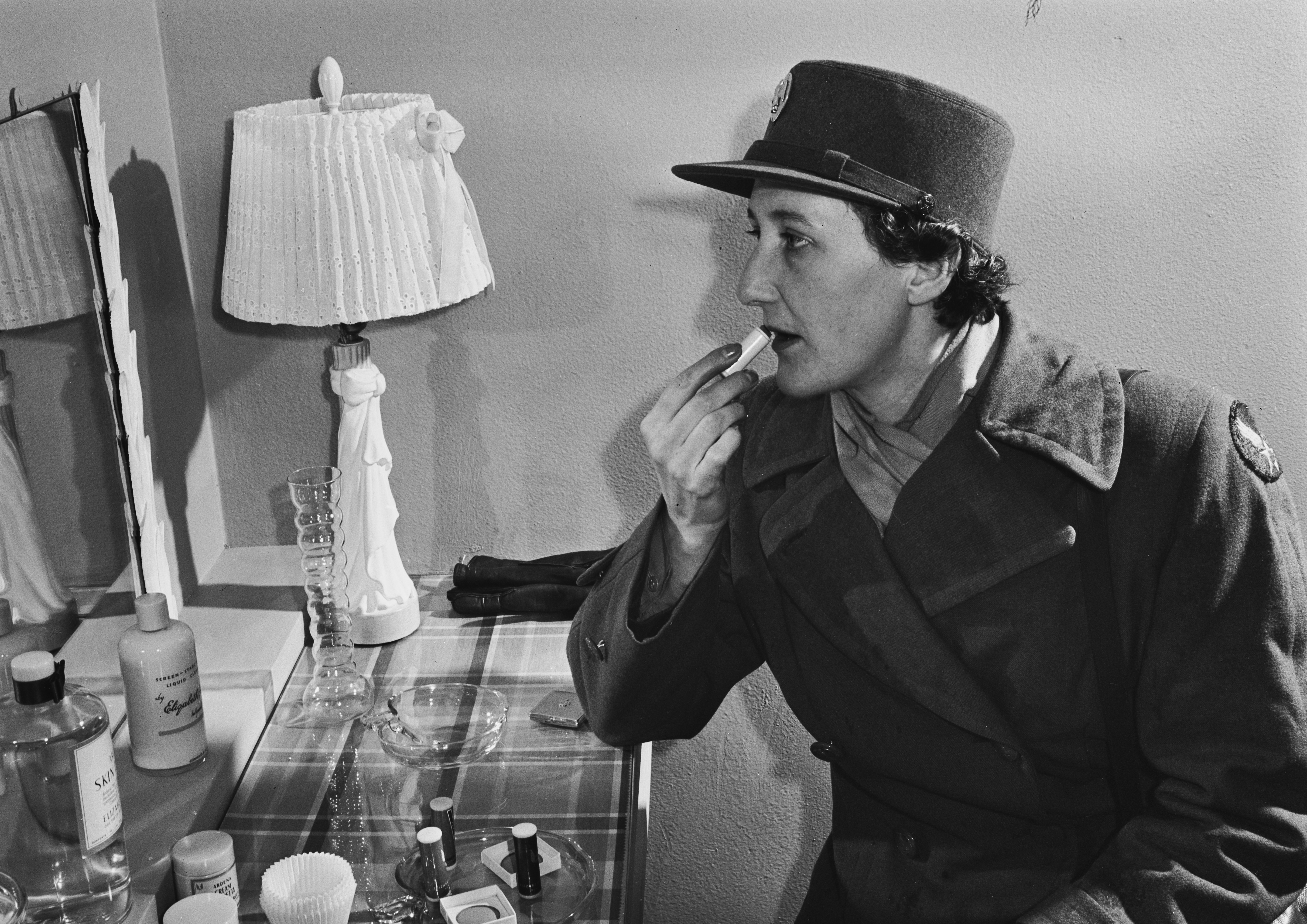 A black and white photograph shows a woman in a uniform, including a dark hat and coat, applying something to her lips. She is seen in profile, looking towards the right side of the frame. To her left, on a patterned surface, are a decorative lamp with a pleated shade, some bottles, and an ashtray.