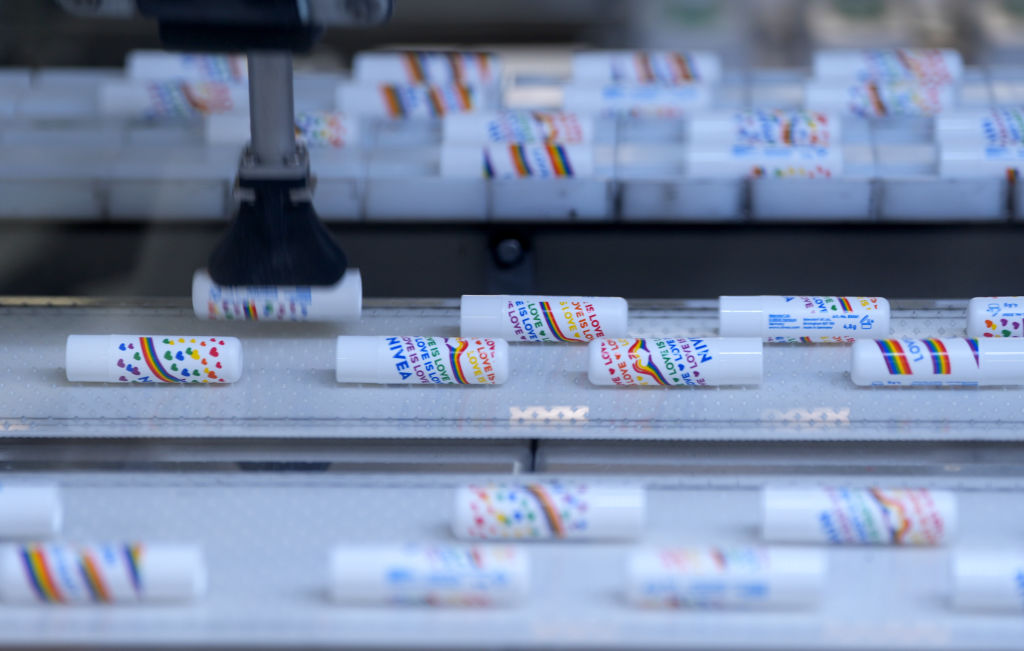 A factory production line shows numerous white tubes of Nivea lip balm, each decorated with a rainbow pattern and the text "LOVE IS LOVE," moving along a conveyor belt. In the foreground, more tubes are visible, while in the background, a robotic arm appears to be positioning or packaging one of the tubes.