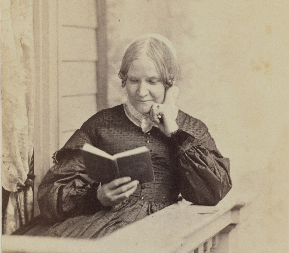 A sepia-toned portrait of Lydia Maria Child, a 19th-century woman with her hair parted in the middle and pulled back. She is sitting outdoors on a porch with a window with lace curtains behind her. She's wearing a dark, textured dress with full sleeves and a white lace collar. She is holding and reading a small book, resting her head thoughtfully on her left hand.