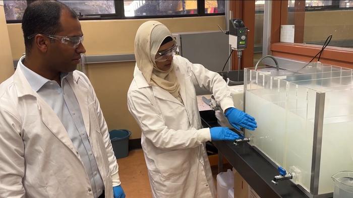 Two scientists in lab coats, safety glasses, and blue gloves examine a clear container of treated wastewater. Dr Biplob Pramanik is pointing at the sample while Dr Nilufa Sultana holds it up for inspection. The photo is taken in a lab at RMIT University.