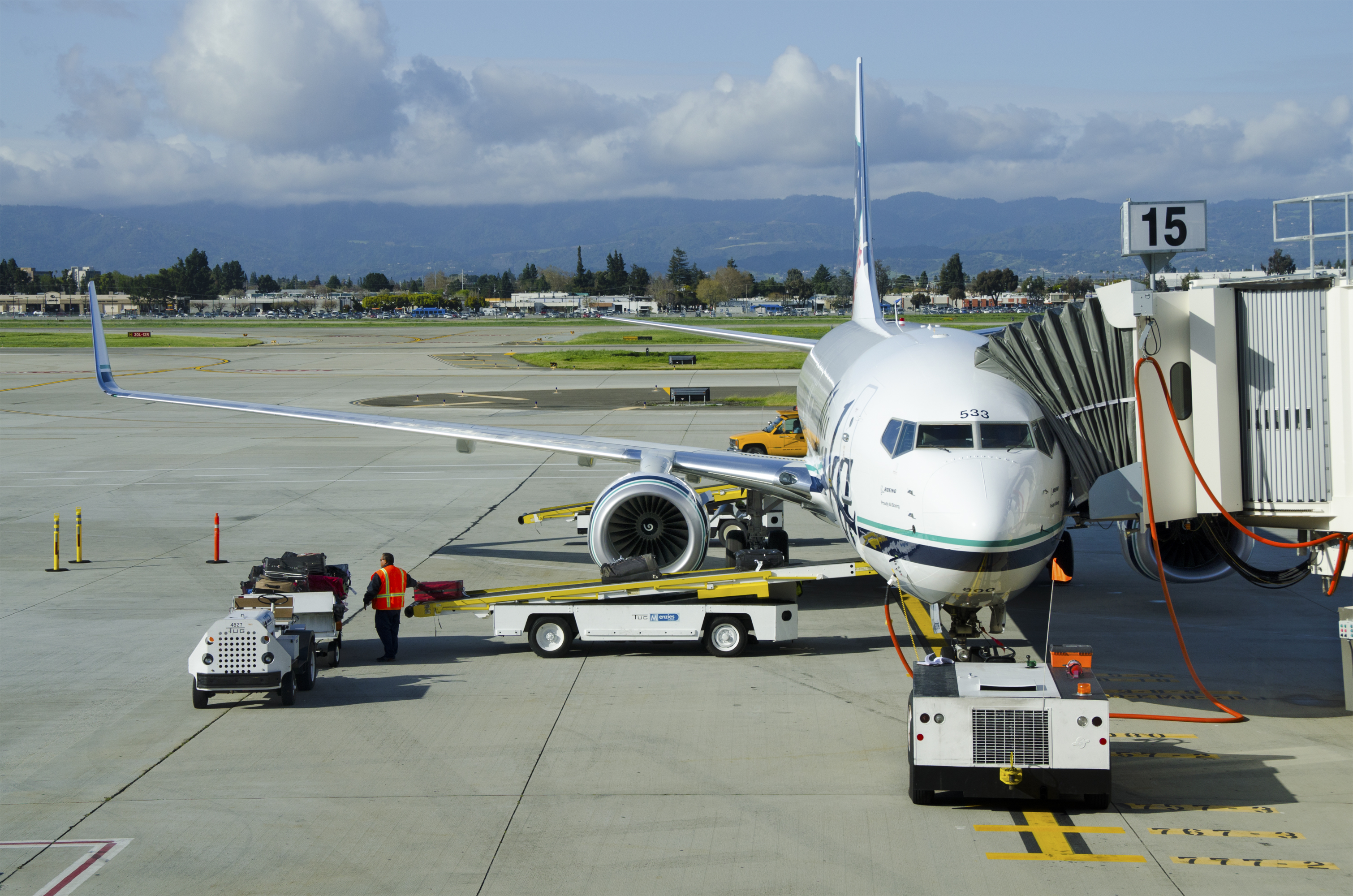 A low-angle, eye-level shot of a Boeing 737-800 airplane from Alaska Airlines parked at a gate on a sunny day. A ground crew worker in a safety vest is visible near the front of the plane, and a baggage conveyor belt is positioned to load luggage into the cargo hold. A jet bridge is connected to the aircraft's front door, and in the distance, low mountains are visible against a partly cloudy sky.
