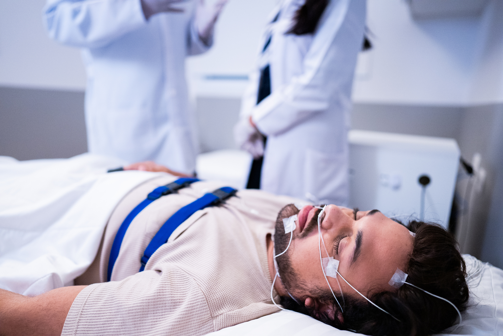 A man lying in a sleep study lab bed with electrodes and monitoring sensors attached to his head, face, and chest. Two medical professionals in white lab coats stand in the background, discussing his condition.