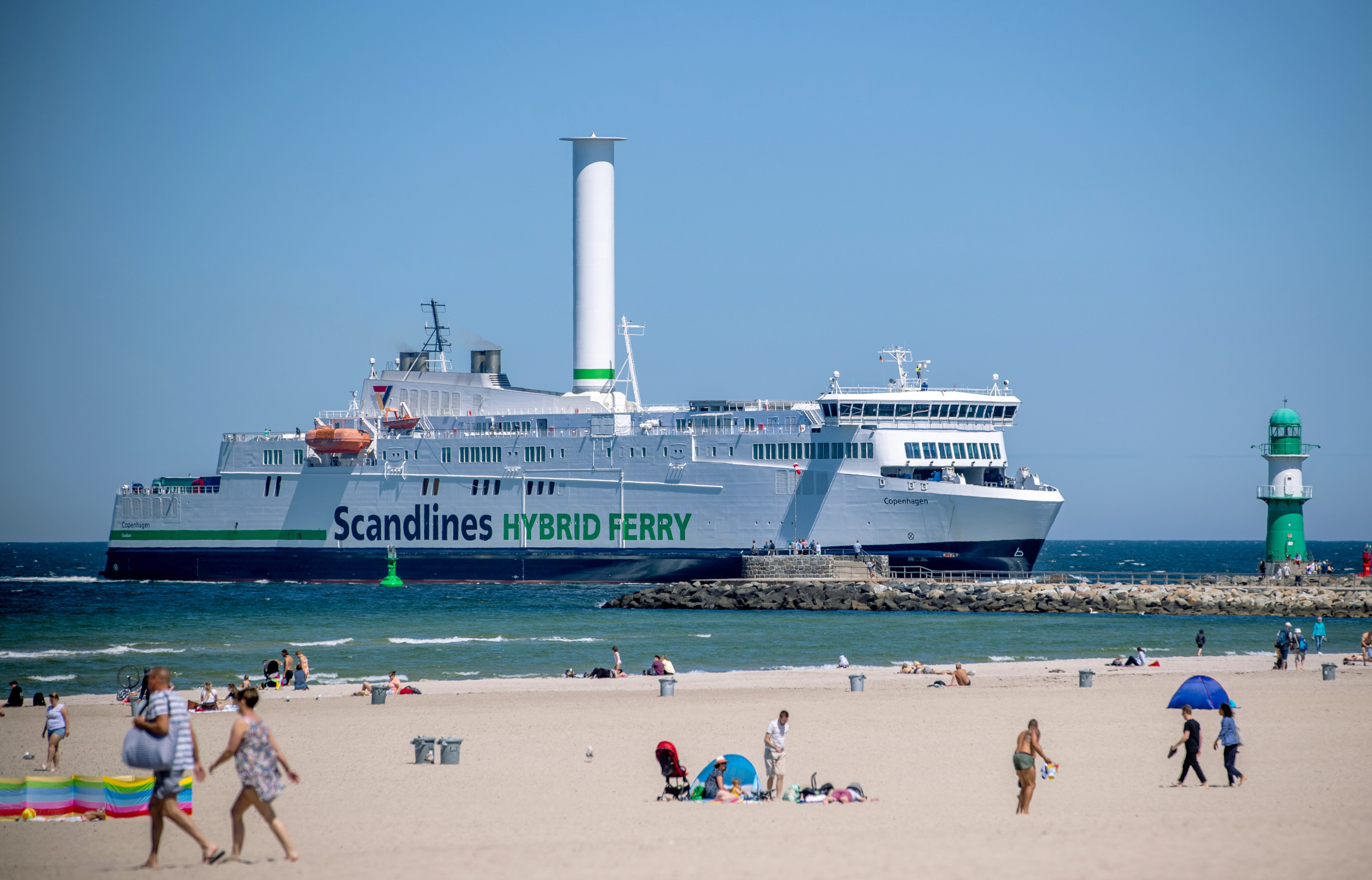 A color photograph shows a large white hybrid ferry with "Scandlines" and "HYBRID FERRY" written on its side. The ferry has a tall, white, spinning rotor sail on its deck. It is moving across a blue sea near a sandy beach with people sunbathing and walking, and a green lighthouse is visible in the distance. The sky is clear and blue.