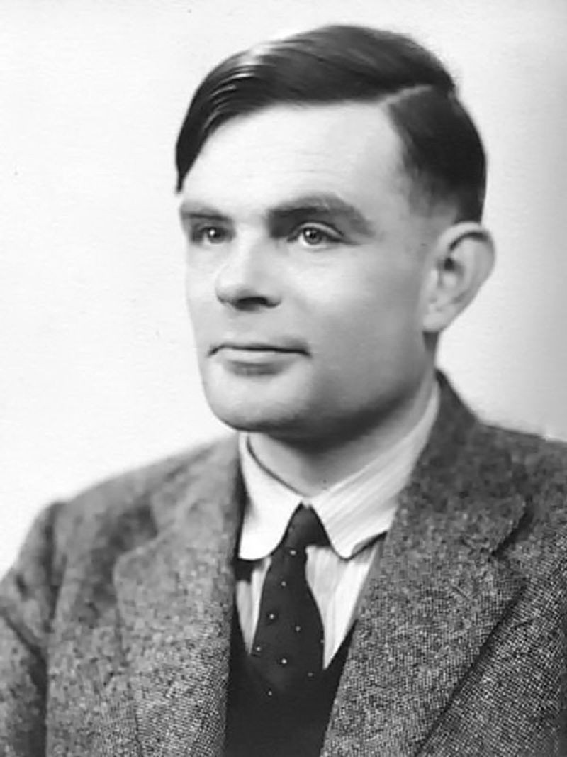 A formal black and white portrait of a young man, Alan Turing, with a calm expression, looking slightly to the side. He has neatly combed dark hair, is wearing a tweed suit jacket, a striped collared shirt, and a polka-dot tie. The background is a plain white or light gray.