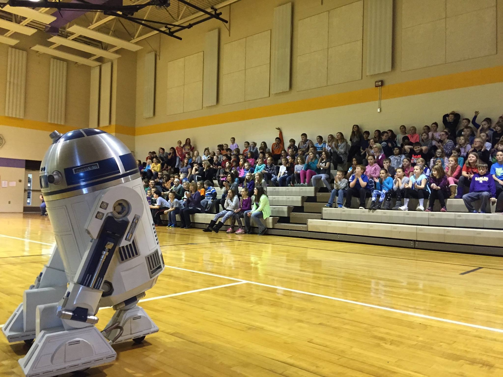 a star wars droid standing in a gymnasium with kids sitting in the stands