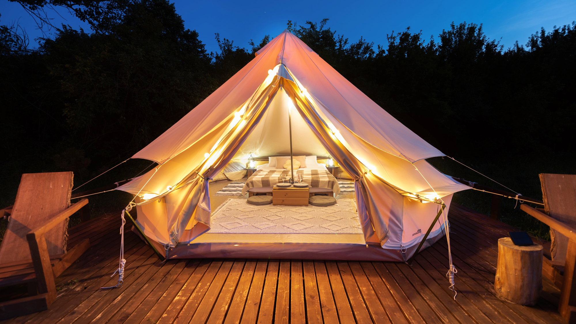 A cozy, lighted glamping tent with its entrance open, revealing a bed with a comforter and pillows, a small table, and a rug inside. The tent is set up on a wooden deck with two chairs and a tree stump table, surrounded by dark trees under a night sky. The interior is lit with a warm, inviting glow.