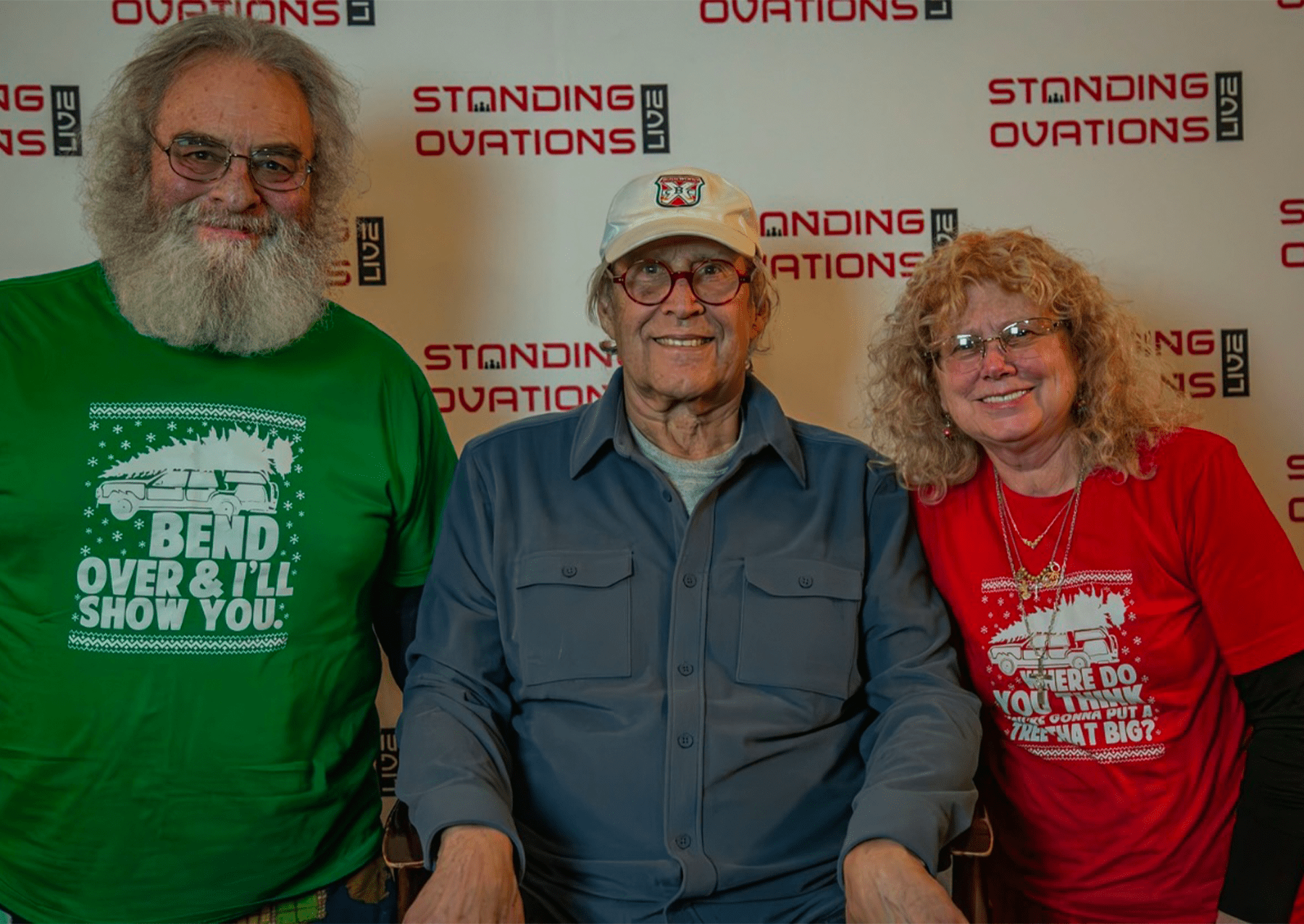 tom dittus wears a green chistmas bacation themed shirt while his wife wears a red one. actor chevy chase is sitting between them