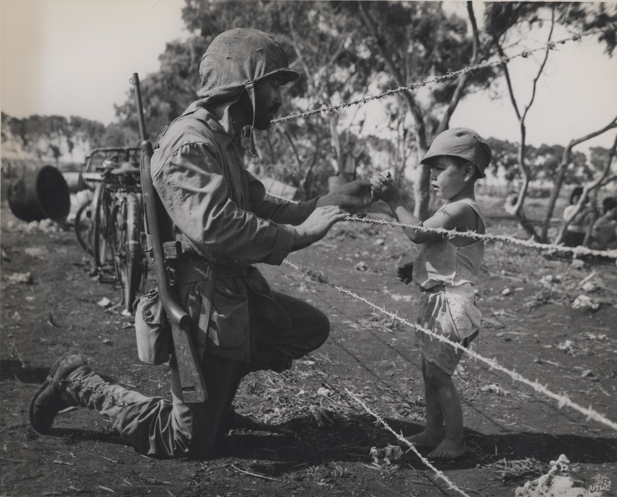 A historical black and white photograph shows a U.S. soldier kneeling on one knee, fully equipped with a helmet and rifle, handing a small object, likely a chocolate bar, to a young boy. The barefoot boy is wearing a cap and light clothes. Three strands of barbed wire separate the two figures, and the background shows a dry, dirt area with trees and scattered debris.