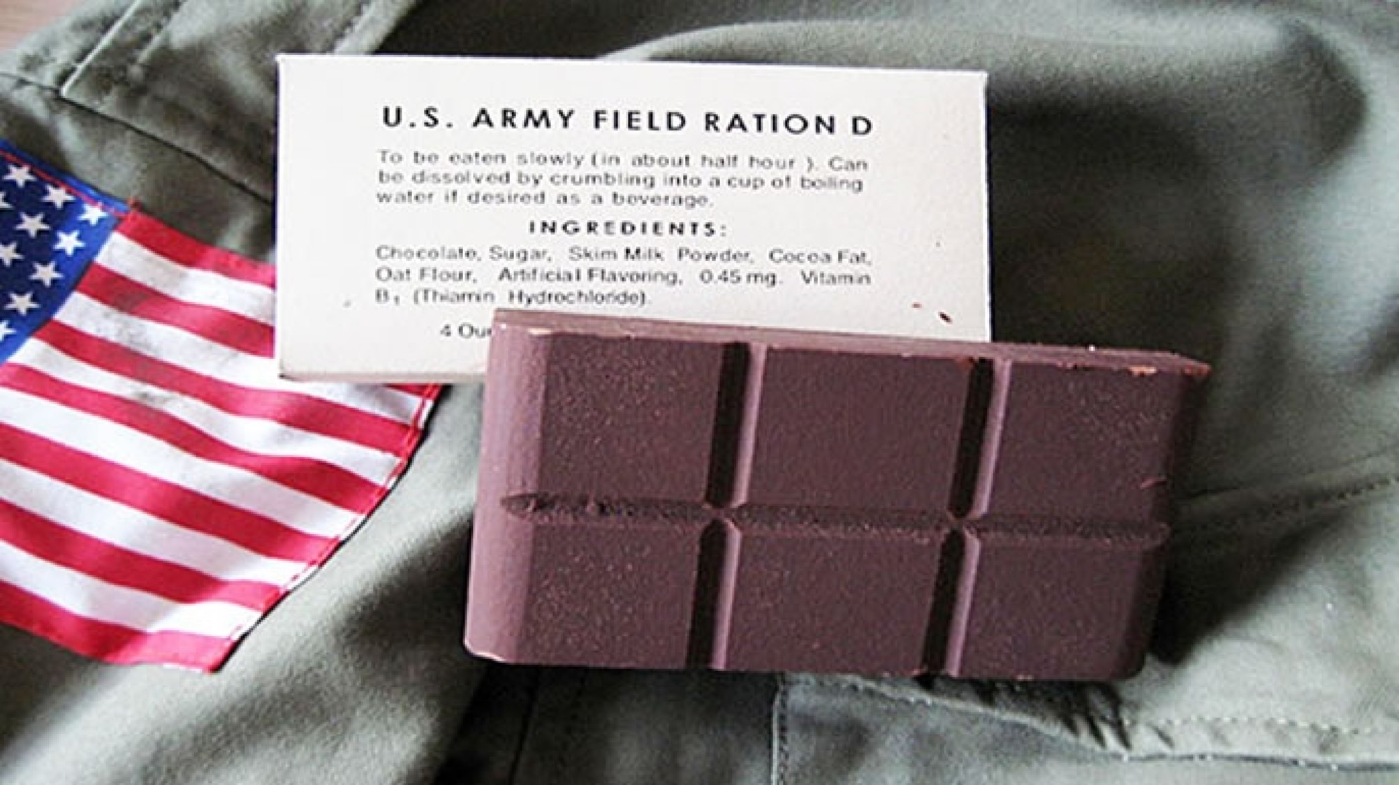A dark brown, thick chocolate bar, divided into four segments, is displayed next to its original white cardboard packaging. The text on the package reads "U.S. ARMY FIELD RATION D" and lists the ingredients. The items are lying on a piece of olive-green military fabric, with a folded corner of an American flag visible in the lower left.