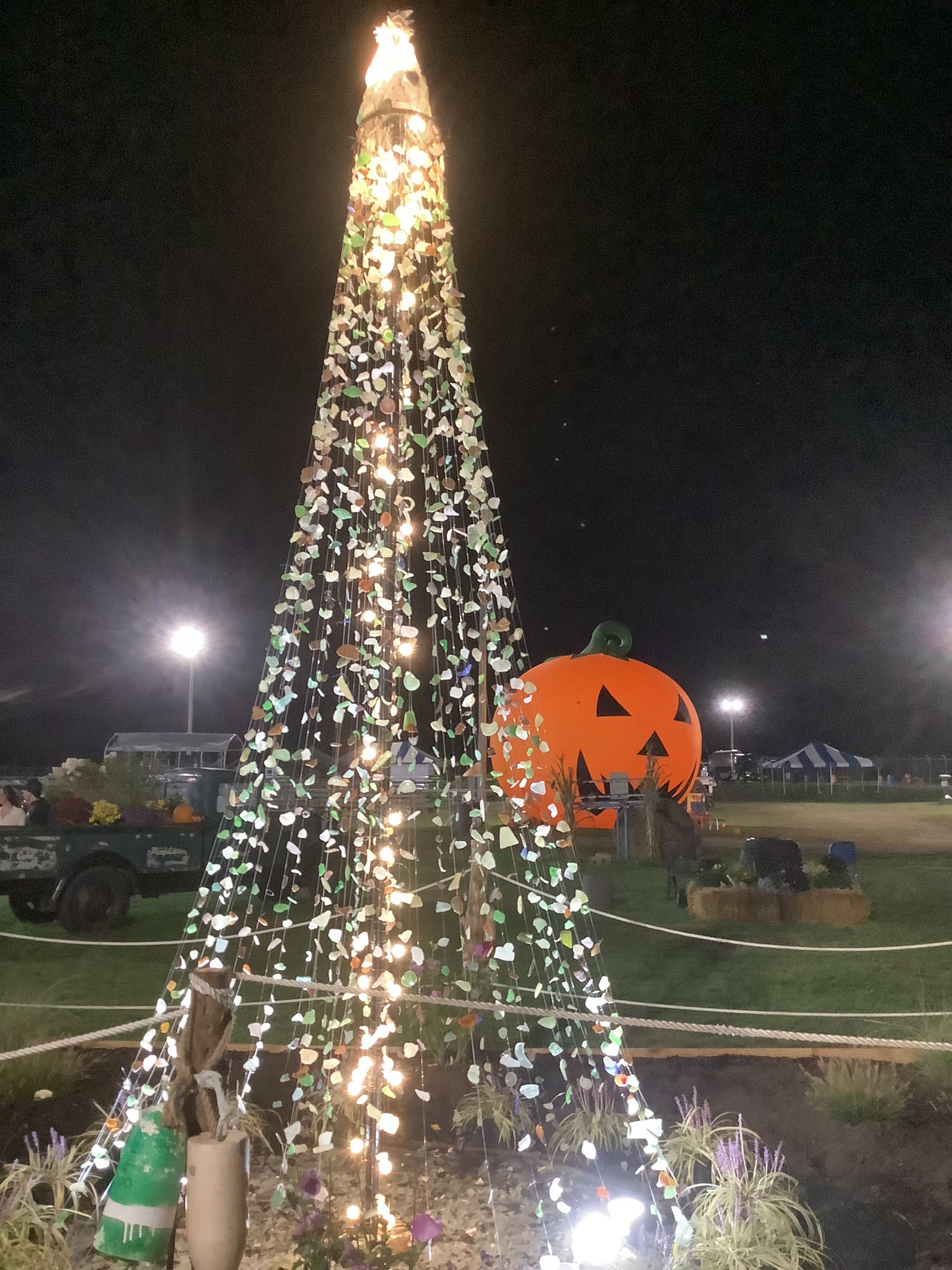 a christmas tree made out of sea glass in a festival with a tractor filled with flowers and an inflateable jack o lantern in the background 