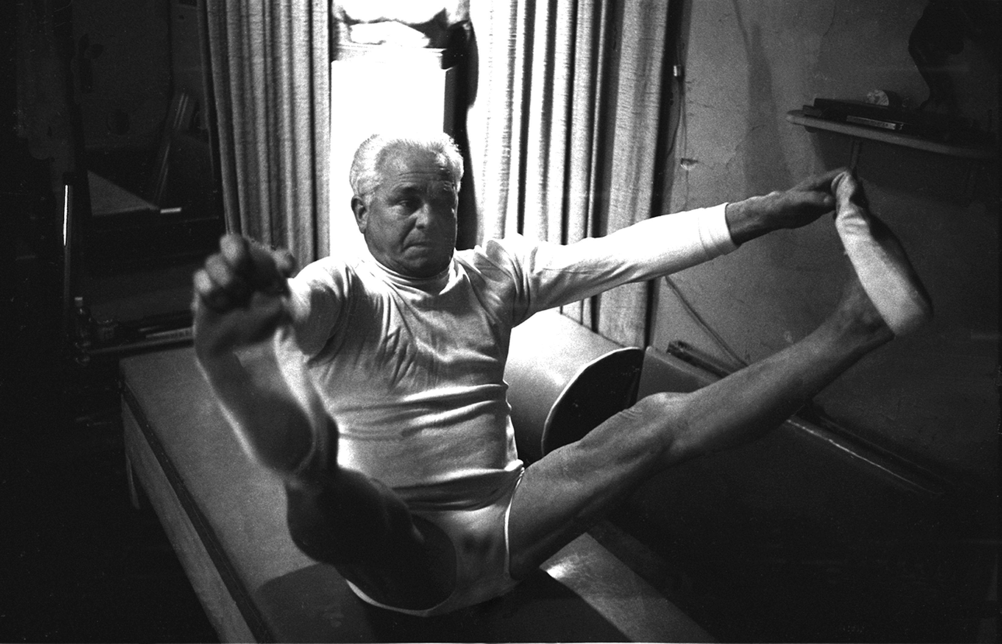 A grainy, black and white archival photo of Joseph Pilates, an older man with white hair, performing a challenging exercise on a mat or piece of equipment. He is wearing a light-colored long-sleeve top and shorts, sitting with his legs extended wide and his arms grasping his feet while keeping his balance. The background is dimly lit with curtains visible behind him.