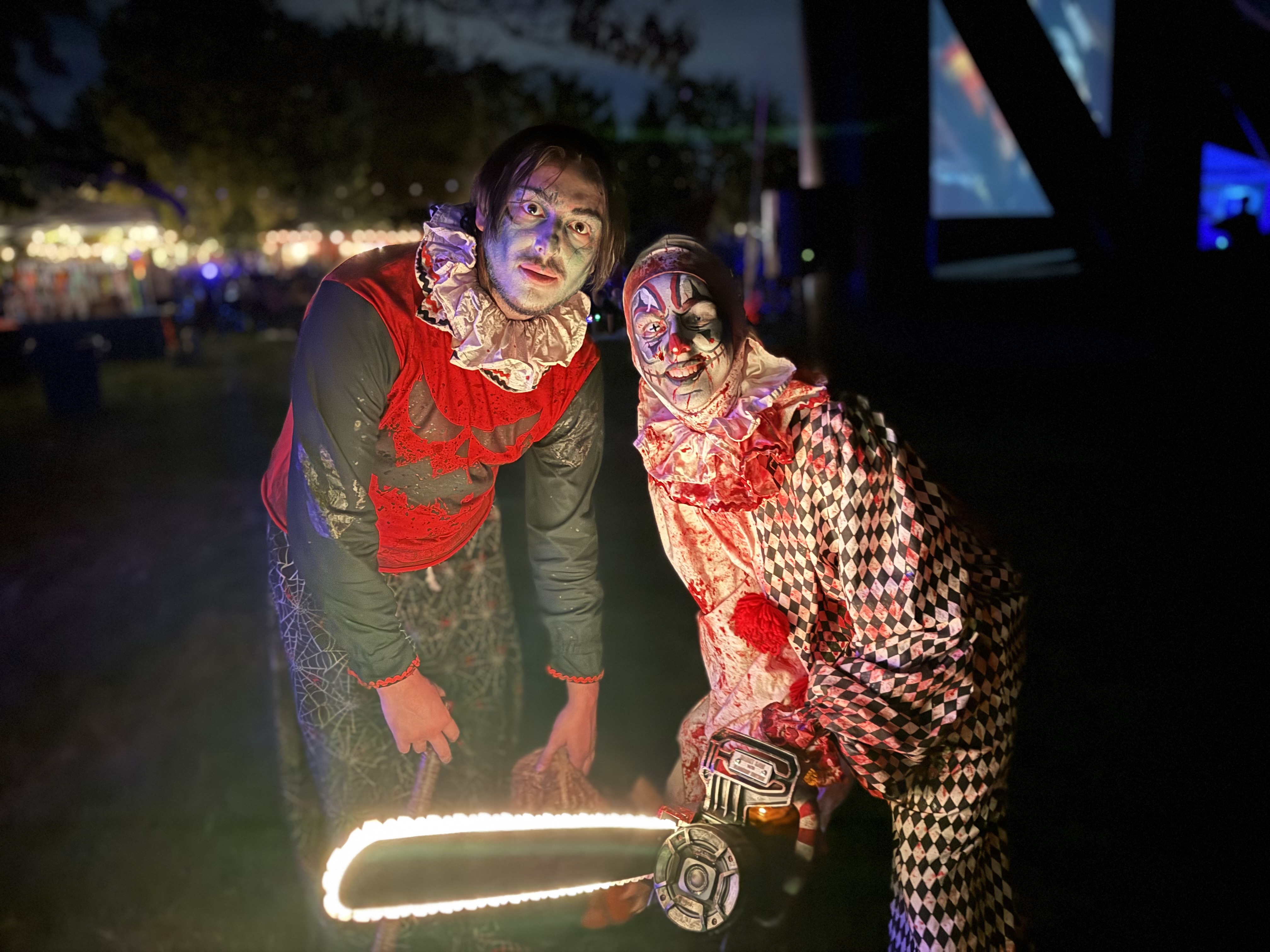 A dark, nighttime photo of two actors in elaborate, frightening clown costumes covered in blood and grime. One clown is wearing a red and black outfit, and the other is wearing a diamond-patterned harlequin suit. They are standing over a prop chainsaw whose blade is glowing with white light, giving them a menacing look. A dark background suggests they are outdoors at a haunted attraction.