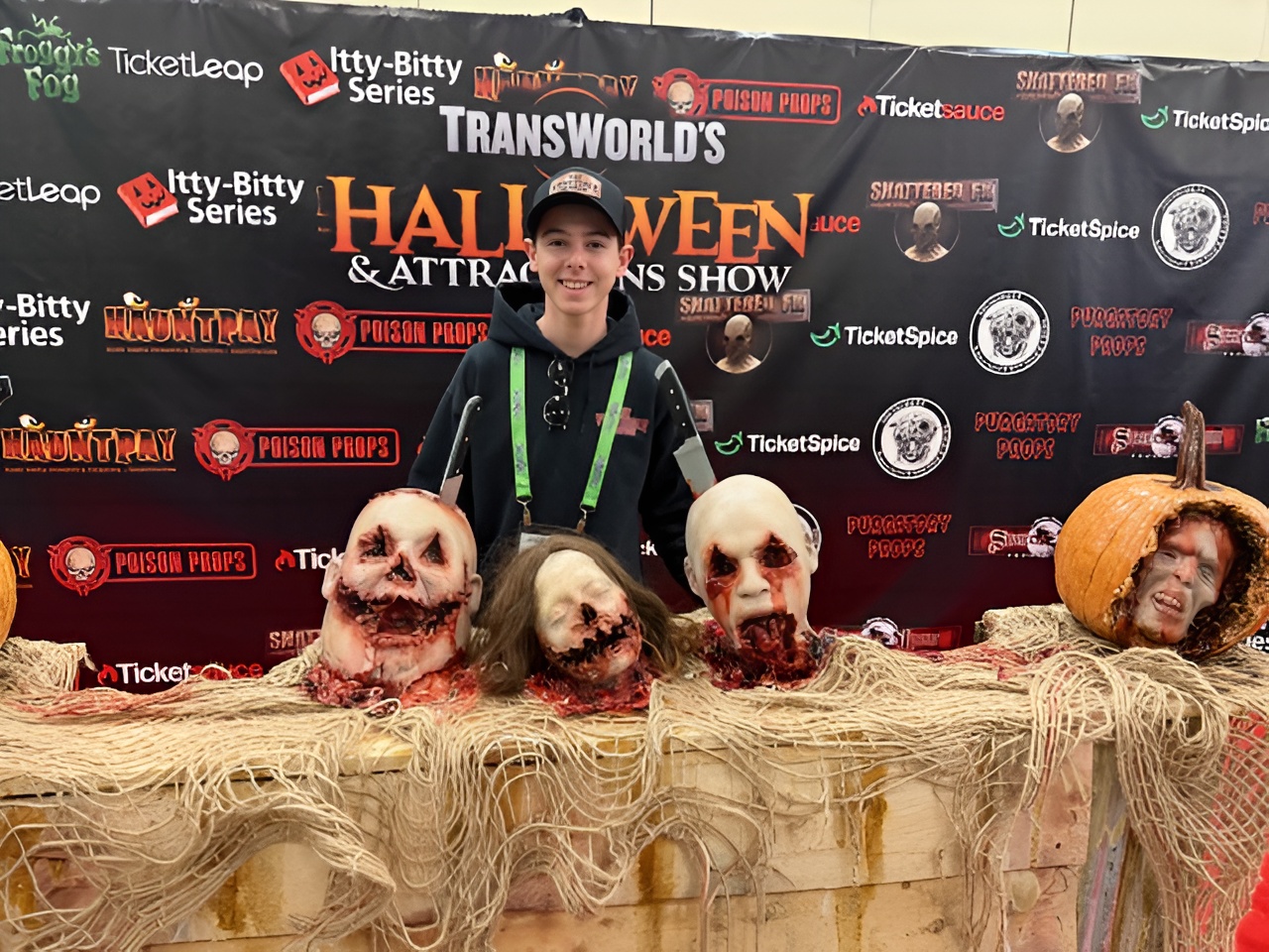 A photo taken at an industry trade show features a smiling young man in a black baseball cap standing behind a table displaying several gruesome haunted house props. The table holds three bloody, severed prop heads with exposed teeth and one carved pumpkin containing a prop head. The background is a black banner for "TRANSWORLD'S HALLOWEEN & ATTRACTIONS SHOW" with various horror-themed logos.