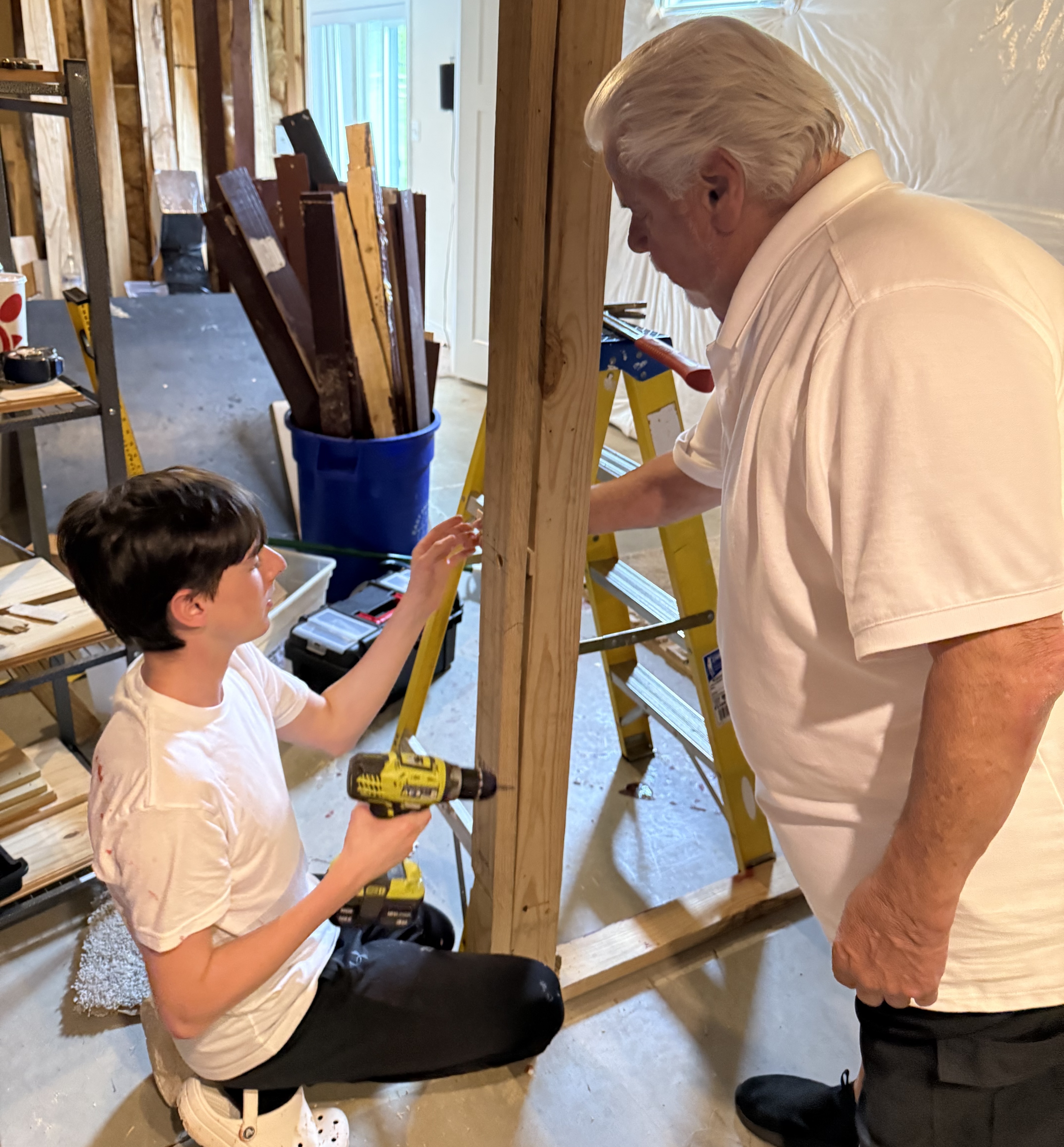 A young boy wearing a white t-shirt and dark pants, kneeling, uses a power drill on a wooden beam, while an older man with white hair, wearing a white polo shirt, stands beside him, supervising or assisting. They are working on a construction project in an unfinished basement, with exposed walls, ductwork, and tools visible in the background.