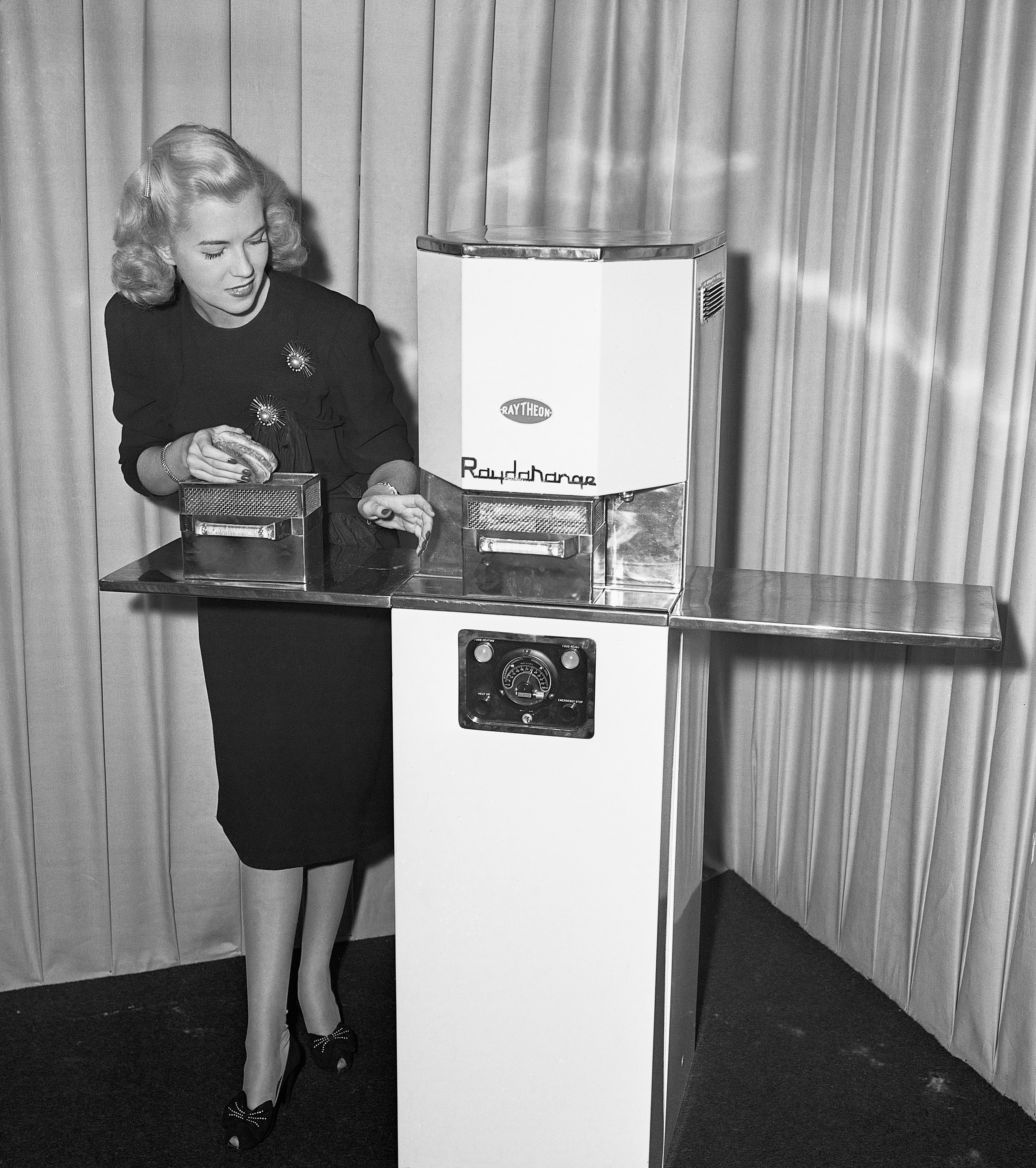 A vintage black and white photograph shows a blonde woman, Jackie Copeland, in a dark dress demonstrating the Raytheon Radarange, one of the earliest commercial microwave ovens, in 1946. The large, industrial-looking appliance is resting on a tall pedestal, and she is holding a small item next to the heating compartment.