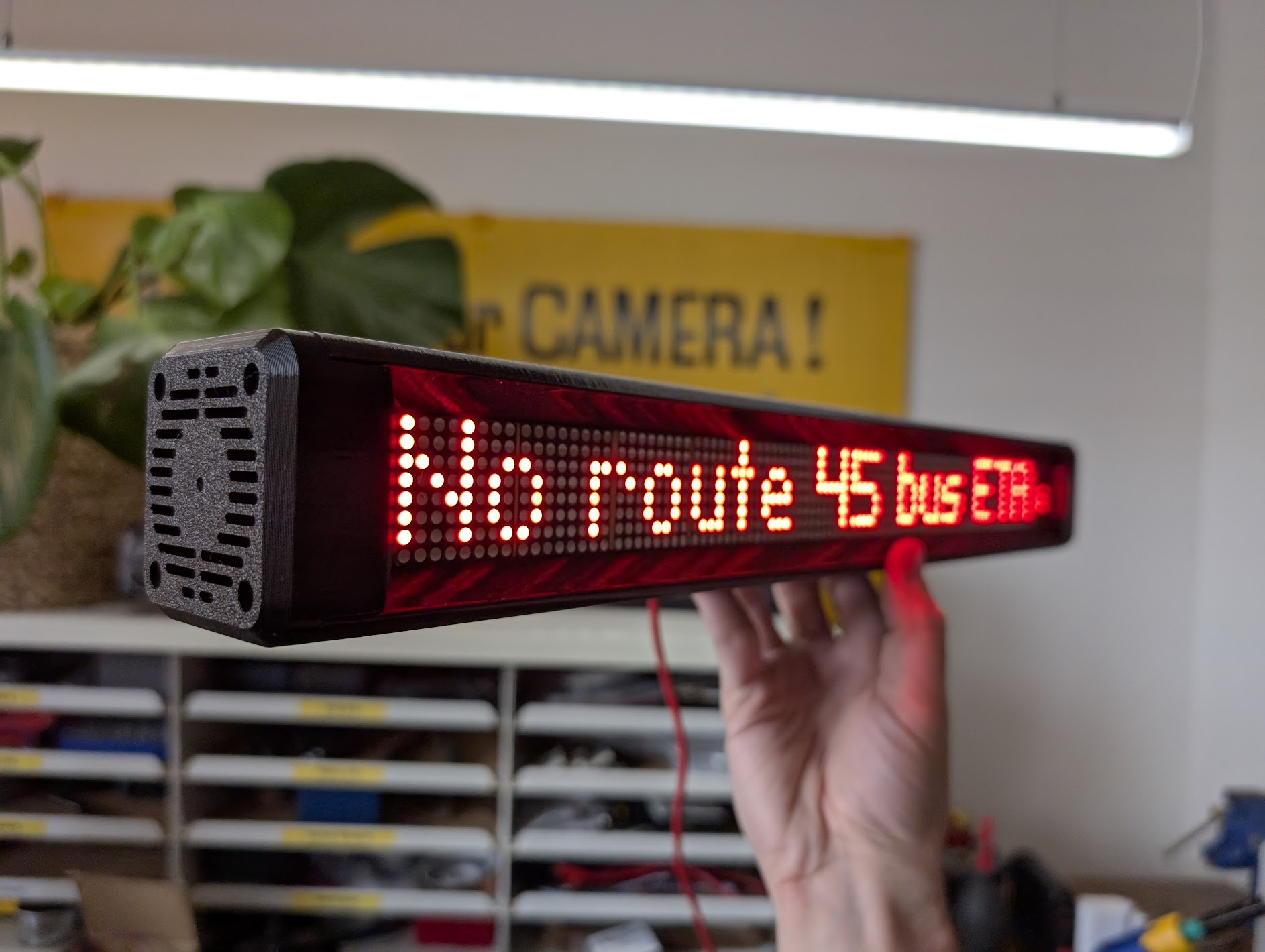 A hand holds a long, black electronic device with a bright red LED display that reads, "No route 45 bus ETA." The device is angled forward, and the background is a blurred office or workshop with a yellow sign reading "CAMERA!" and shelving filled with small drawers.