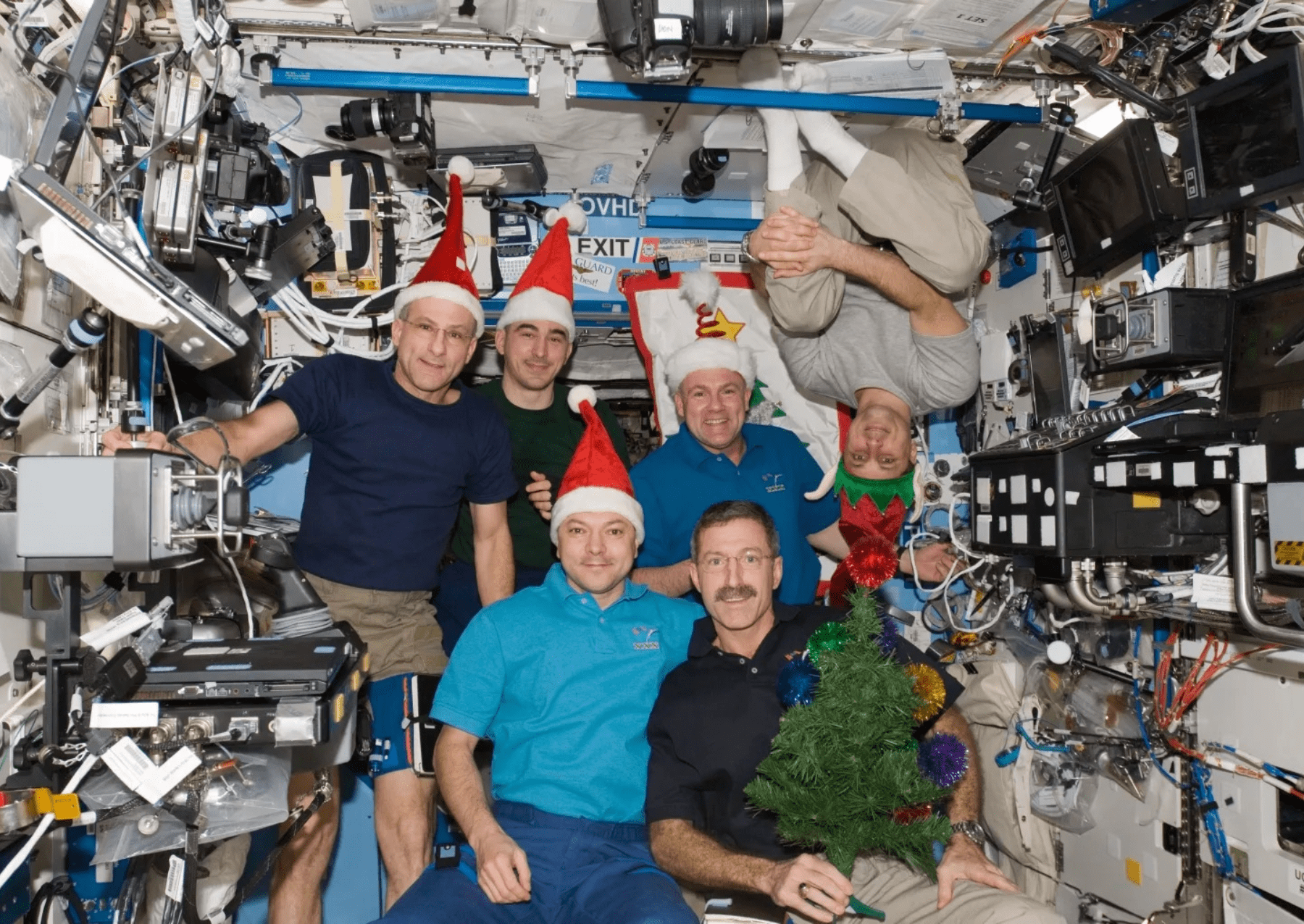 astronauts posing with santa hats and a christmas tree. one wearing an elf hat is upside down