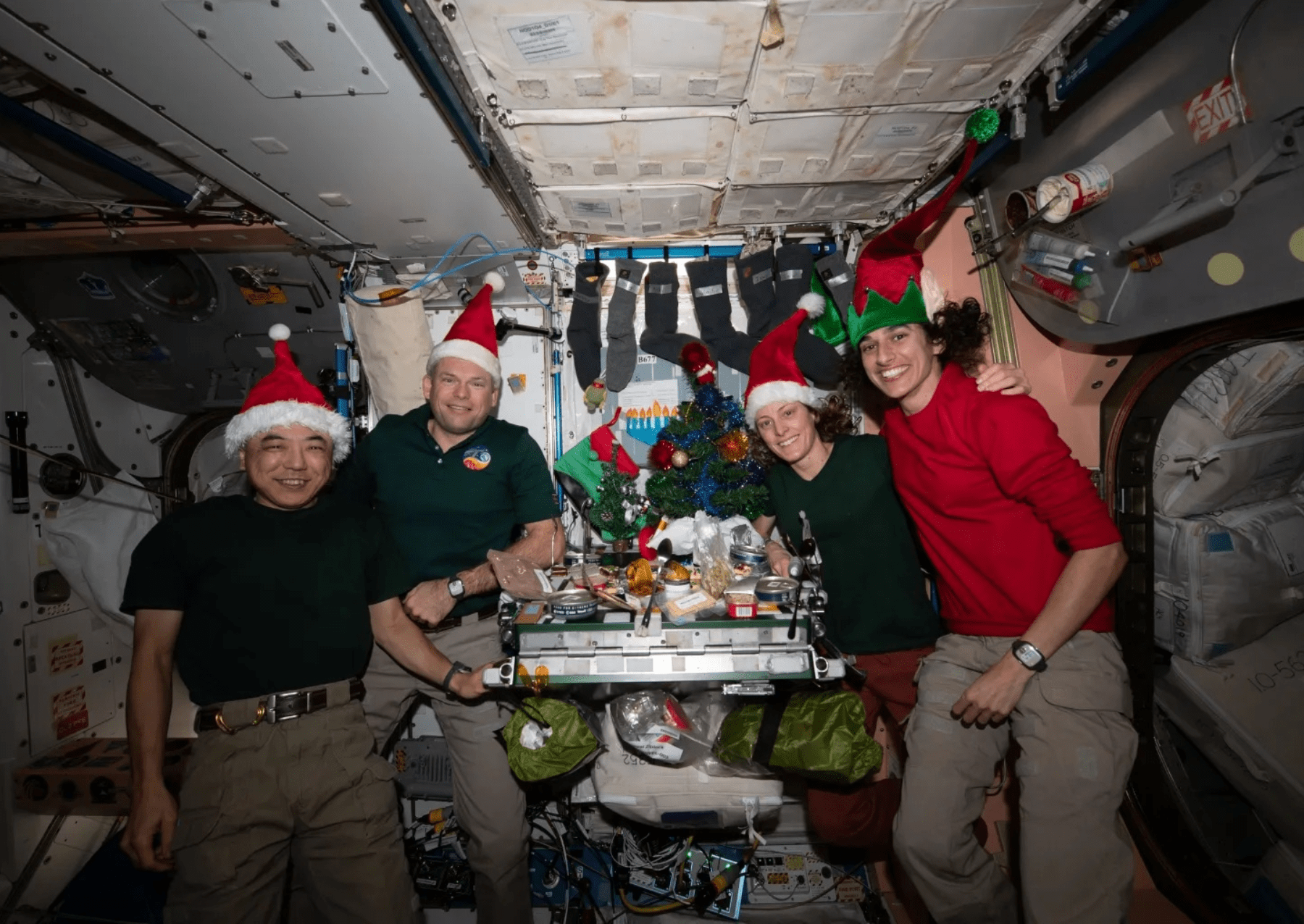 four astronauts gather around a table wearing santa hats