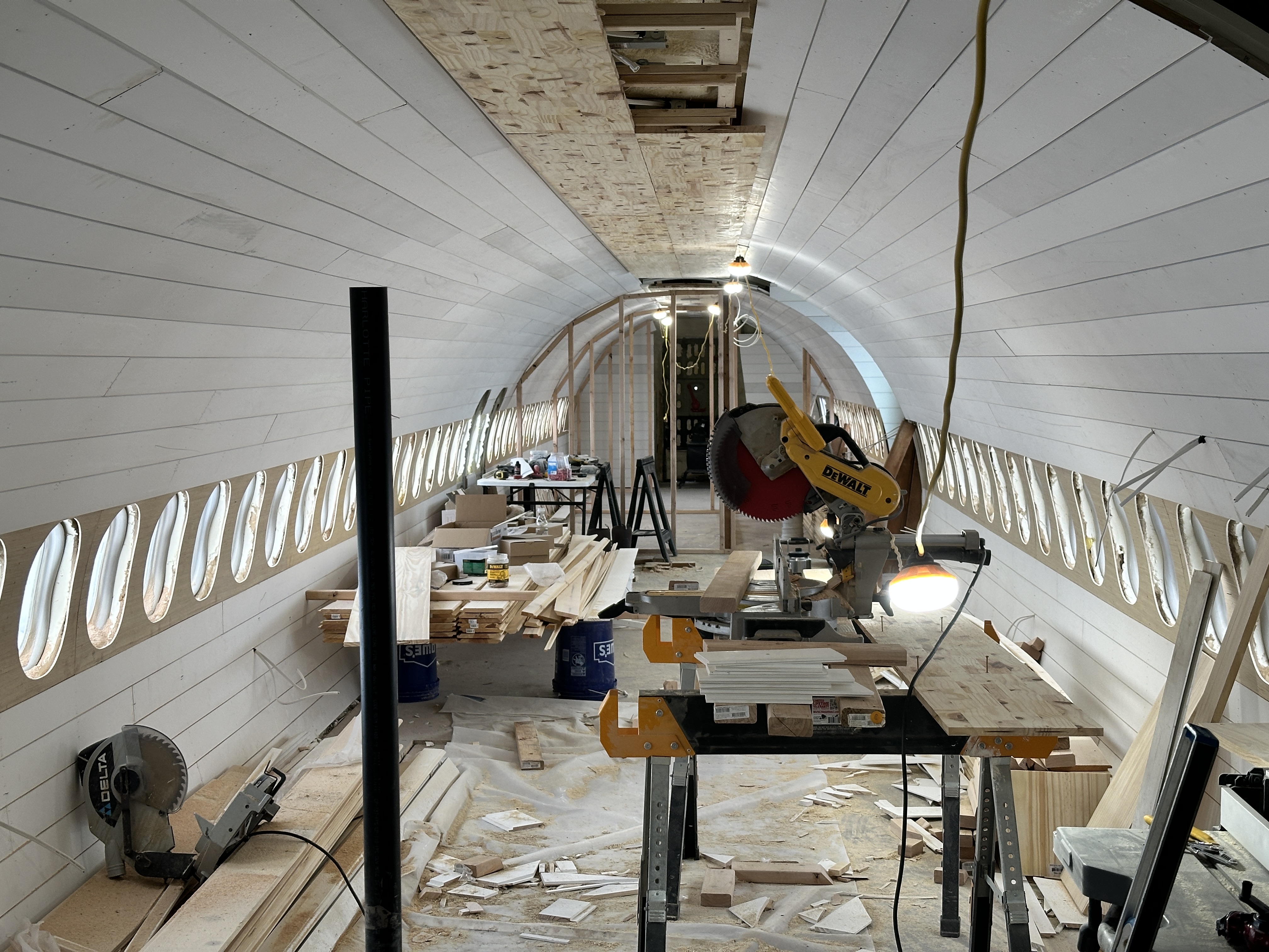 A view of the interior of a Douglas DC-9 airplane fuselage undergoing renovation. The walls are covered in white shiplap paneling, exposing the rows of oval windows and the curved ceiling. The space is filled with construction debris, lumber, and power tools like miter saws, showing the project is in the early stages of conversion into living quarters.