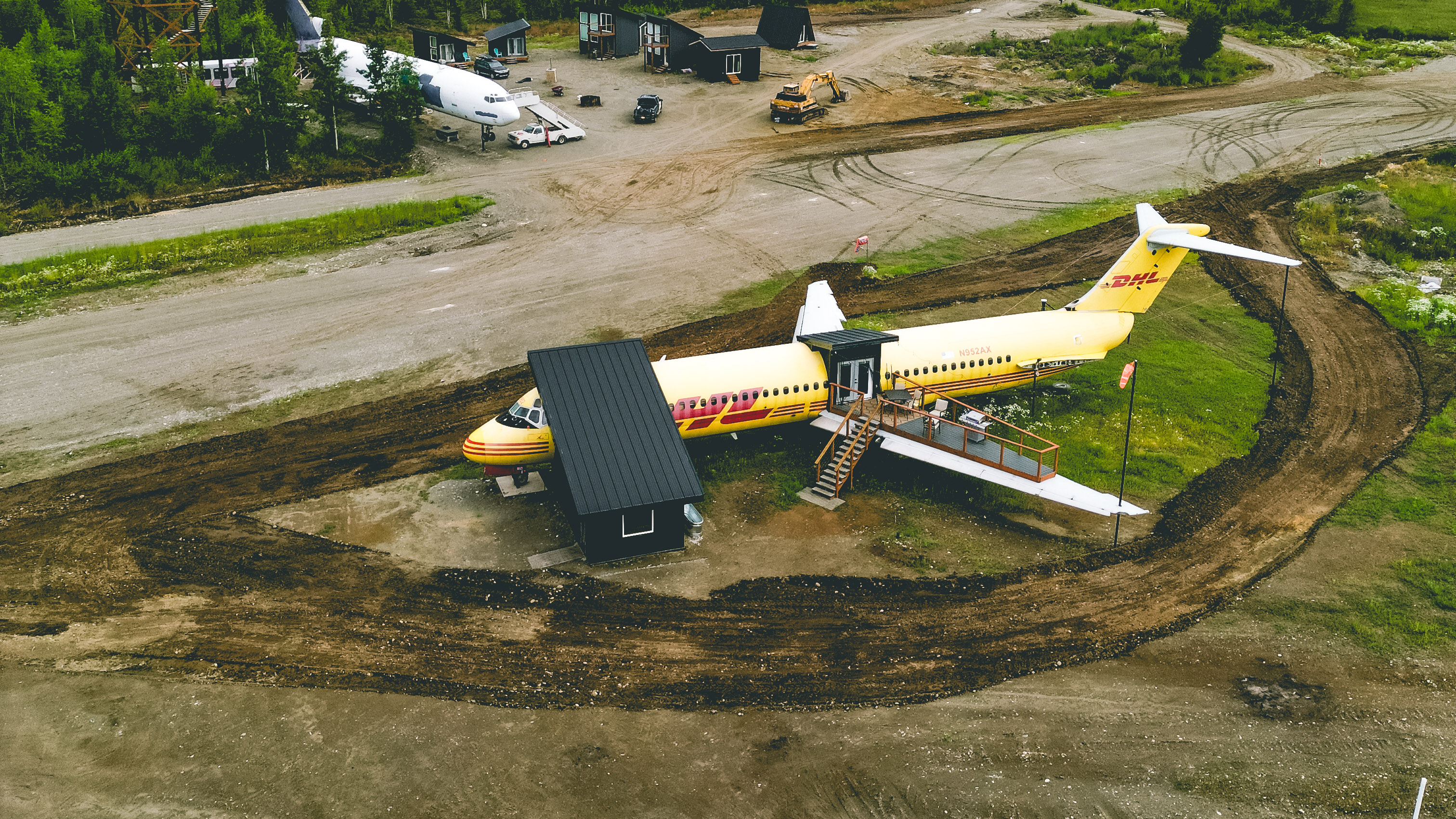 An aerial view of a bright yellow airplane fuselage, previously a DHL cargo plane, that has been converted into unique living quarters. The plane is sitting in a dirt and grassy field, with a set of wooden stairs and a small, modern entryway built onto the wing and fuselage. Several other small, modern cabins or tiny homes are visible in the background among the trees.