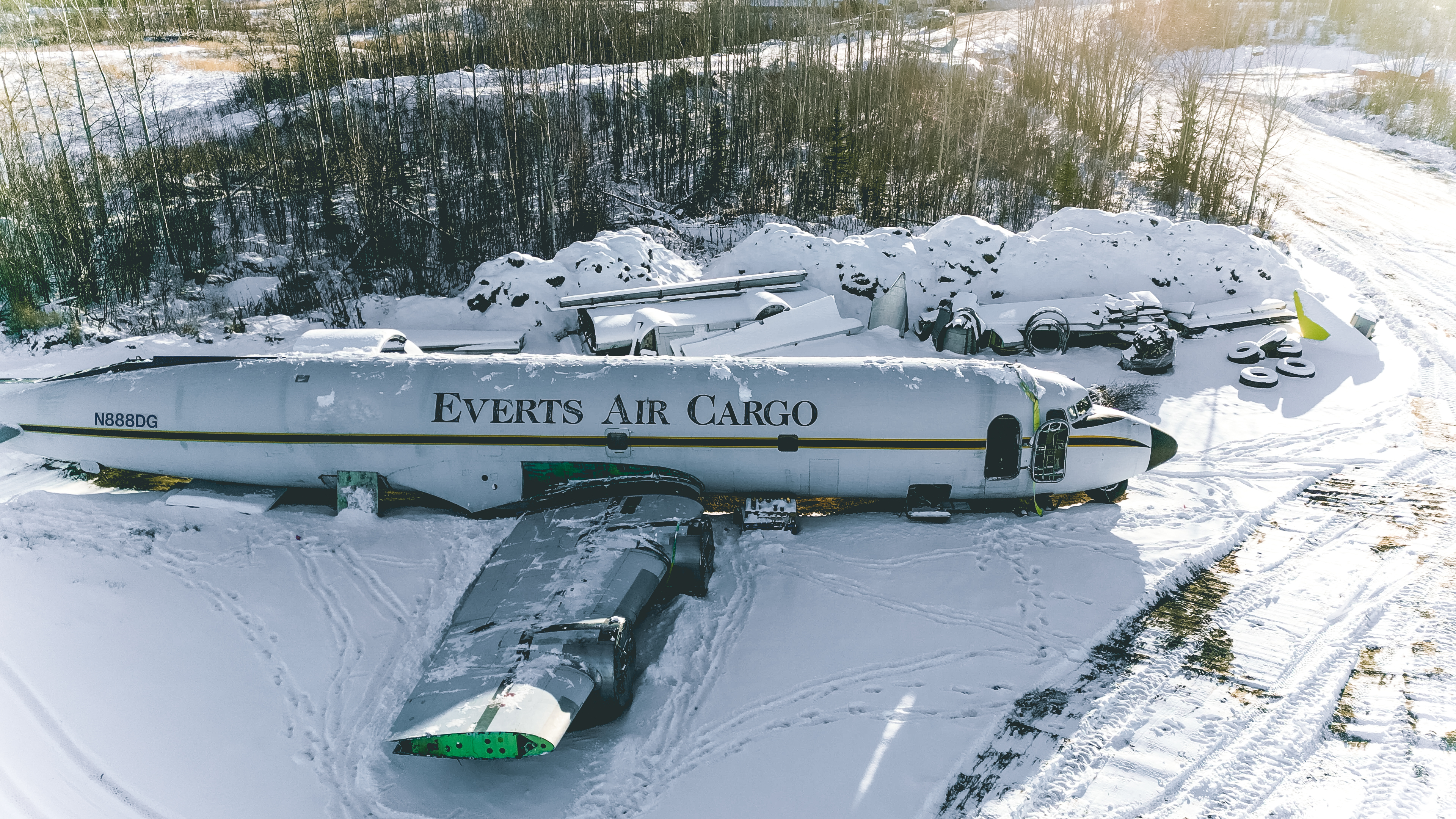 An aerial, wide-angle photo of a large, white Douglas DC-6 propeller airplane fuselage lying disabled in a snowy, open field. The plane has "EVERTS AIR CARGO" painted on its side in gold lettering, and its detached wing rests next to the body. The surrounding area is covered in snow, with trees and brush visible in the background.