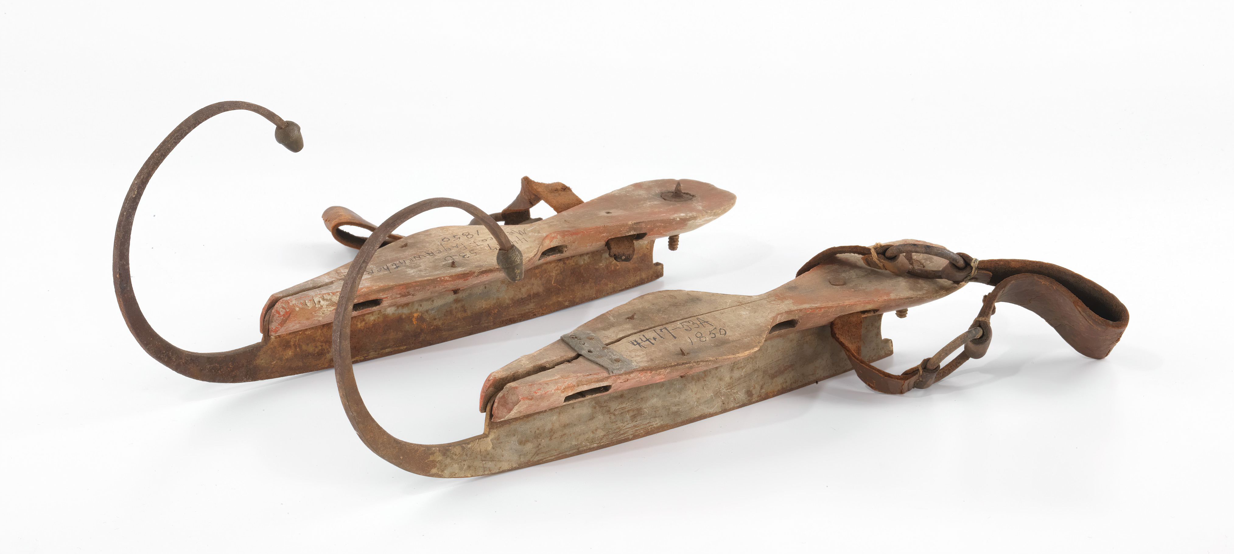 A studio photograph against a white background featuring a pair of antique American ice skates from the mid-nineteenth century. The skates consist of worn wooden foot platforms with rusty metal blades underneath that curve upward into dramatic, large curls at the front. Dark, weathered leather straps are attached to the wooden bases for securing them to a wearer's boots.